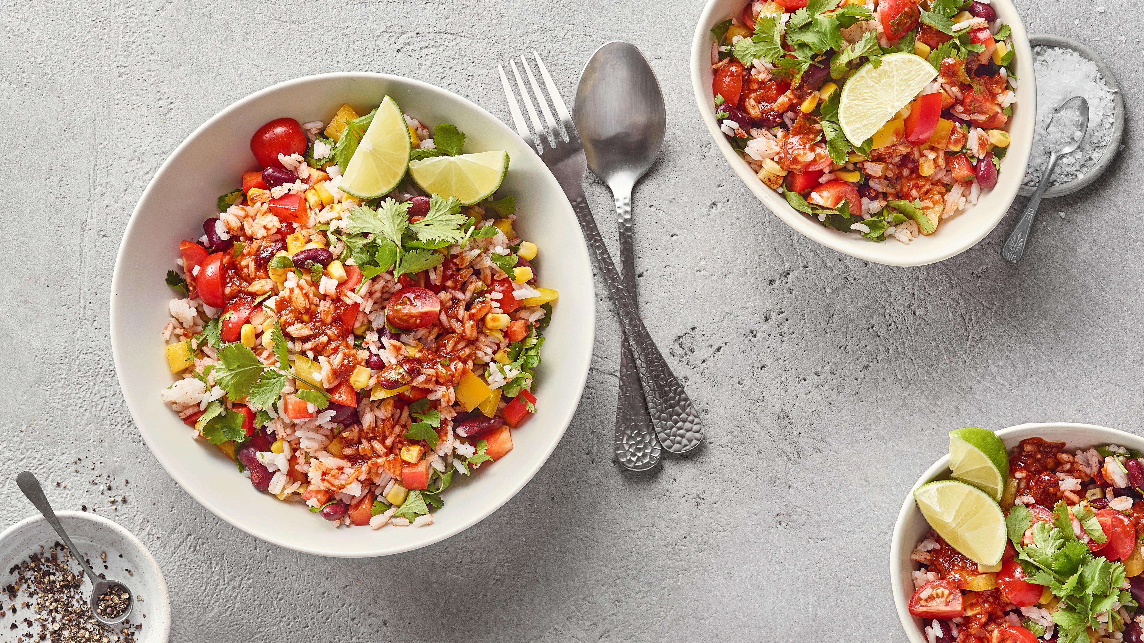 Eine Schüssel mit Reis, roten Bohnen, Mais, Paprikastücken, Tomatenhälften, frischen Kräutern und Limettenspalten.