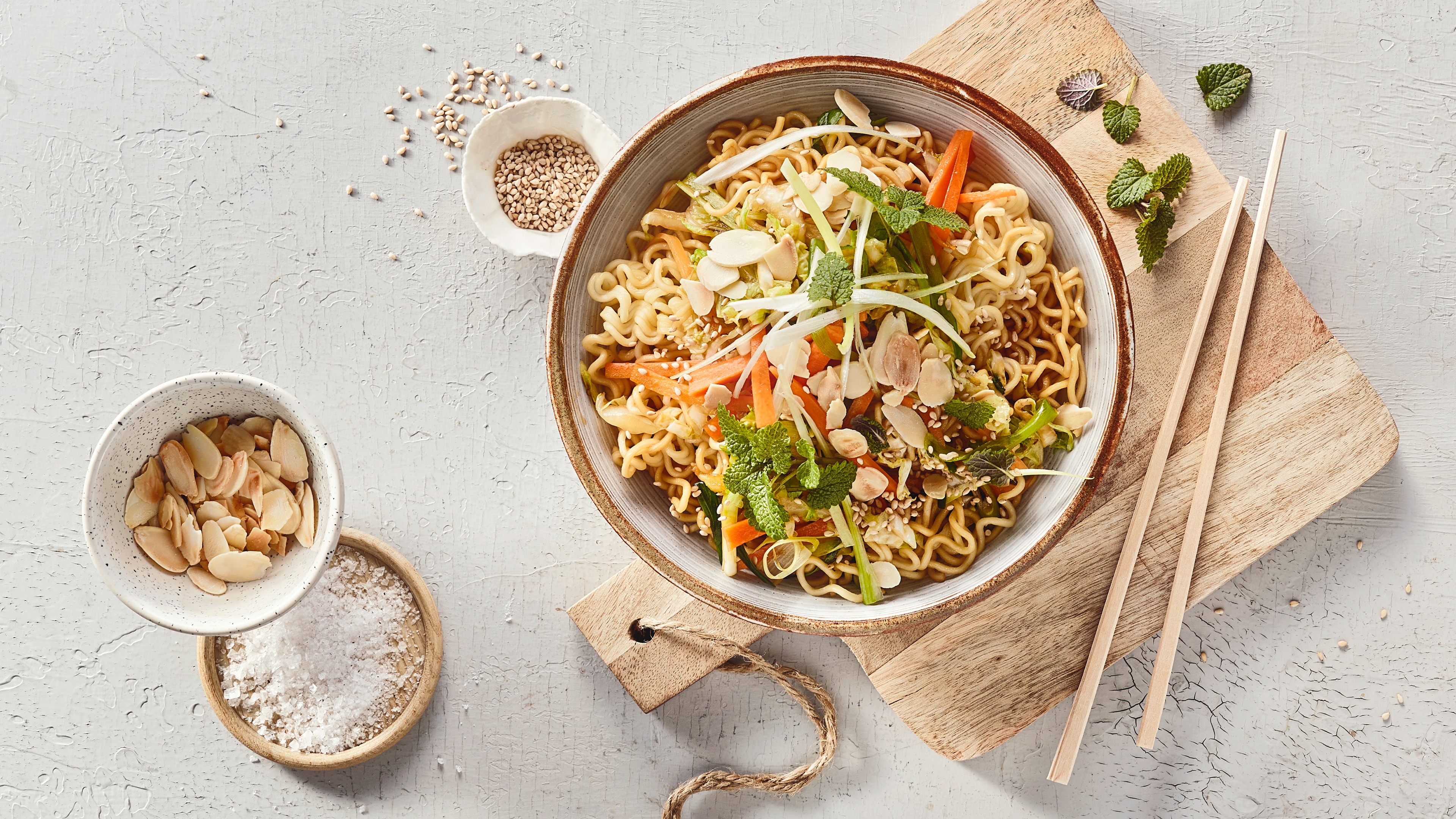 Eine Schüssel mit Nudeln, darauf orange Streifen, frische Kräuter, weiße Ringe, helle Scheiben und kleine Körner.