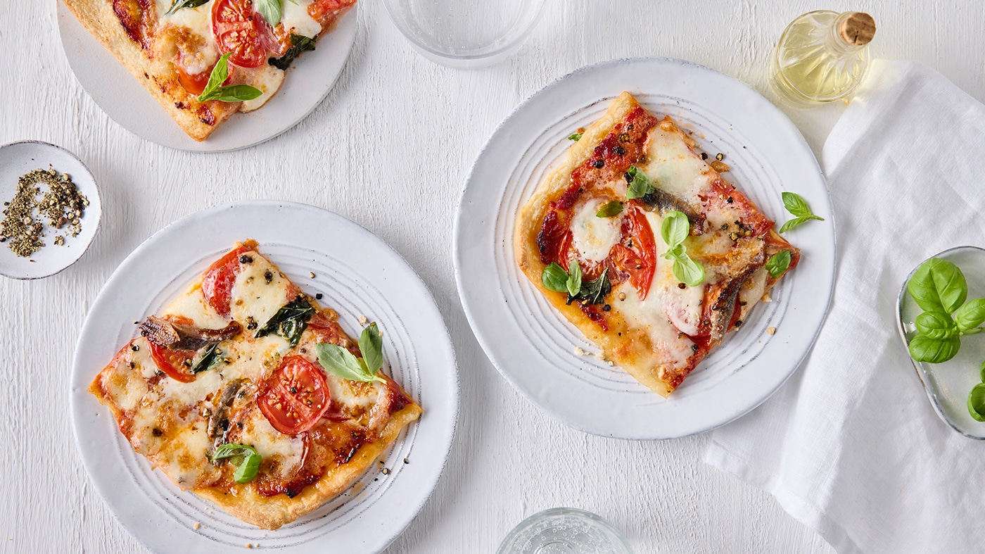 Drei runde, weiße Teller mit jeweils einem belegtem Pizzastück. Zwischen den Tellern stehen Olivenöl sowie Schalen mit Gewürzen und Petersilie.