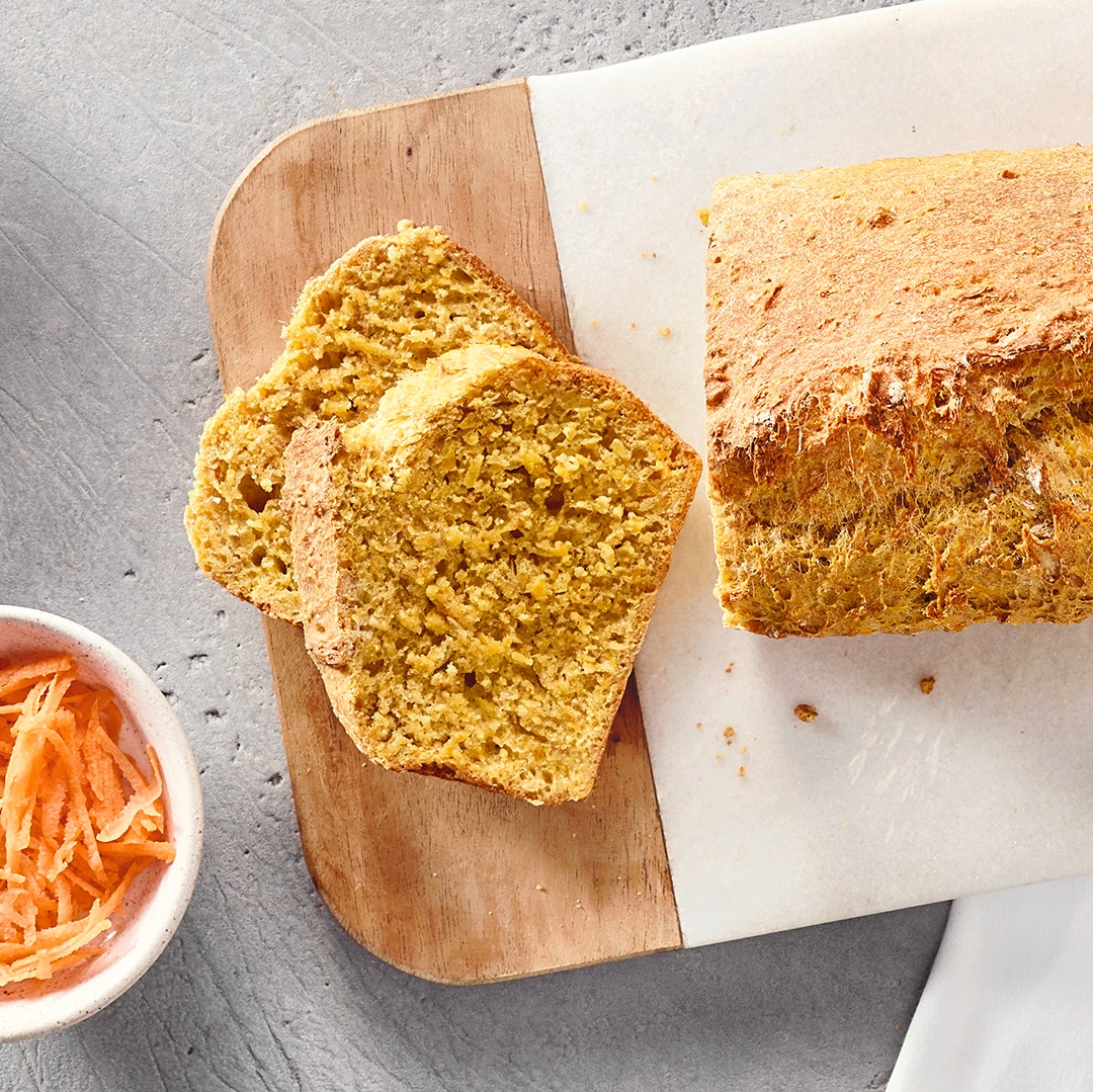 Ein Brett mit einem ganzen Laib und zwei Scheiben hellbraunen Brots, daneben eine Schale mit feinen orangefarbenen Streifen.