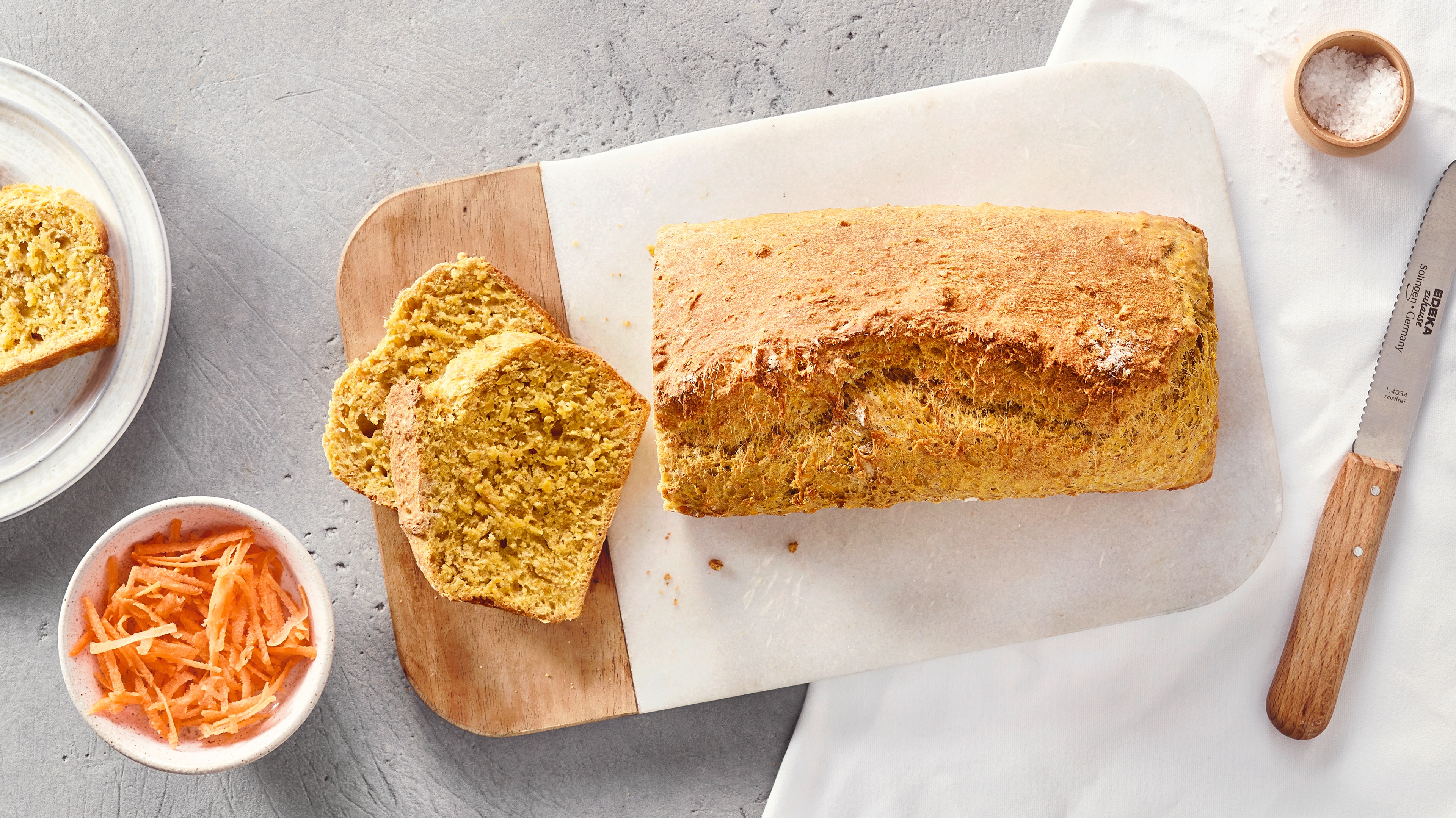 Ein Brett mit einem ganzen Laib und zwei Scheiben hellbraunen Brots, daneben eine Schale mit feinen orangefarbenen Streifen.