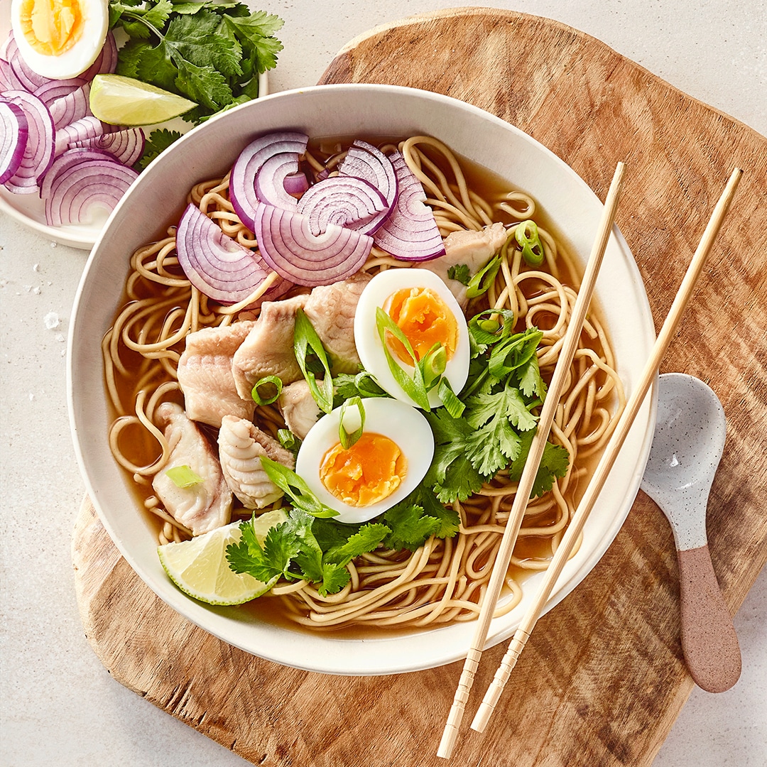 Teller auf Holzbrett mit Nudeln, Zwiebeln, Fischstücken und 2 Eierhälften in brauner Flüssigkeit.