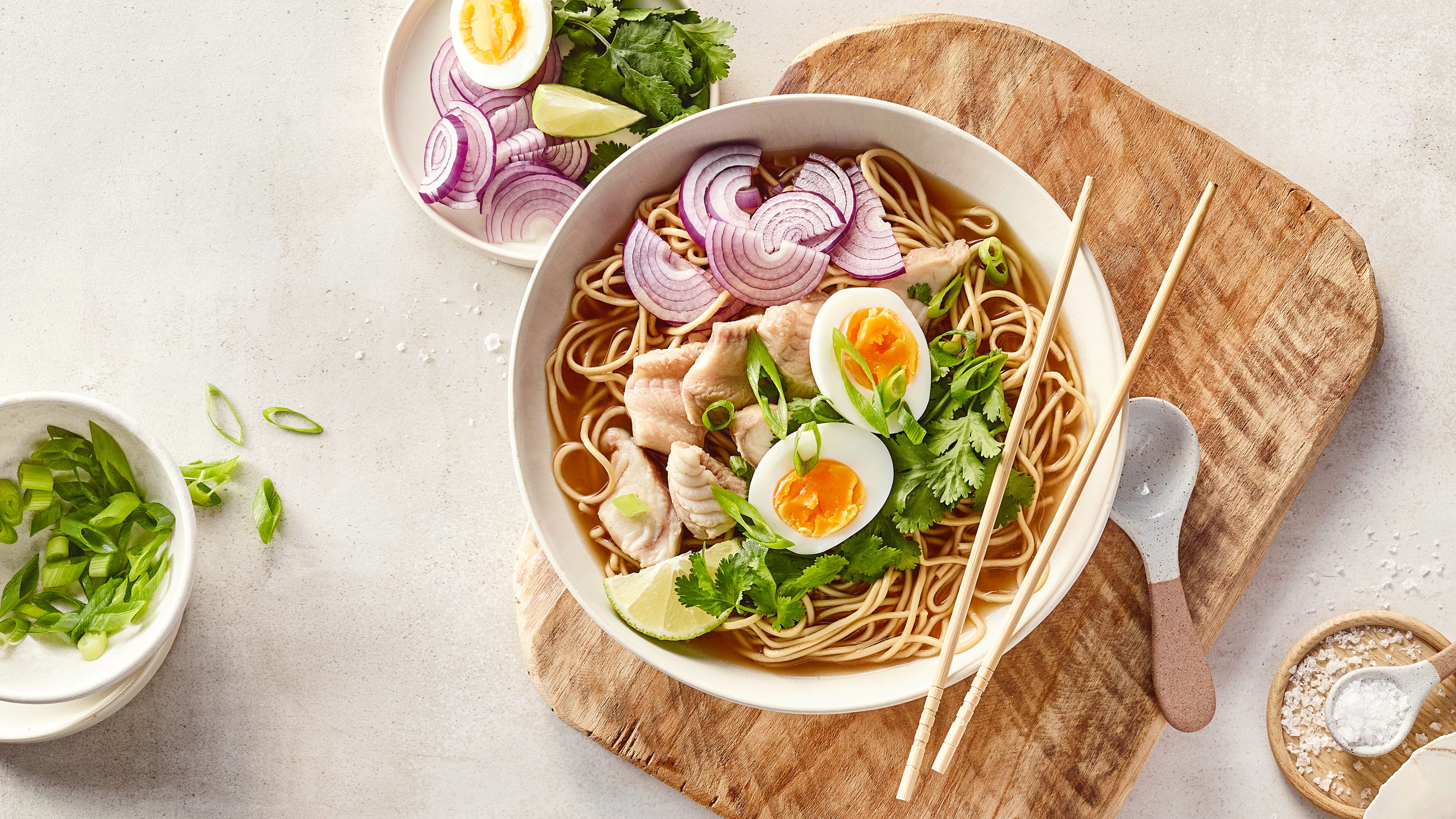 Teller auf Holzbrett mit Nudeln, Zwiebeln, Fischstücken und 2 Eierhälften in brauner Flüssigkeit.