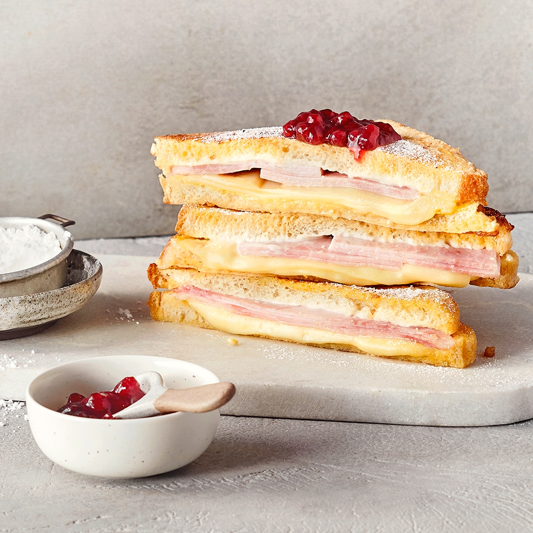 Ein Brett mit drei getoasteten Sandwiches, gefüllt mit Schinken und geschmolzenem Käse, garniert mit rotem Gelee..