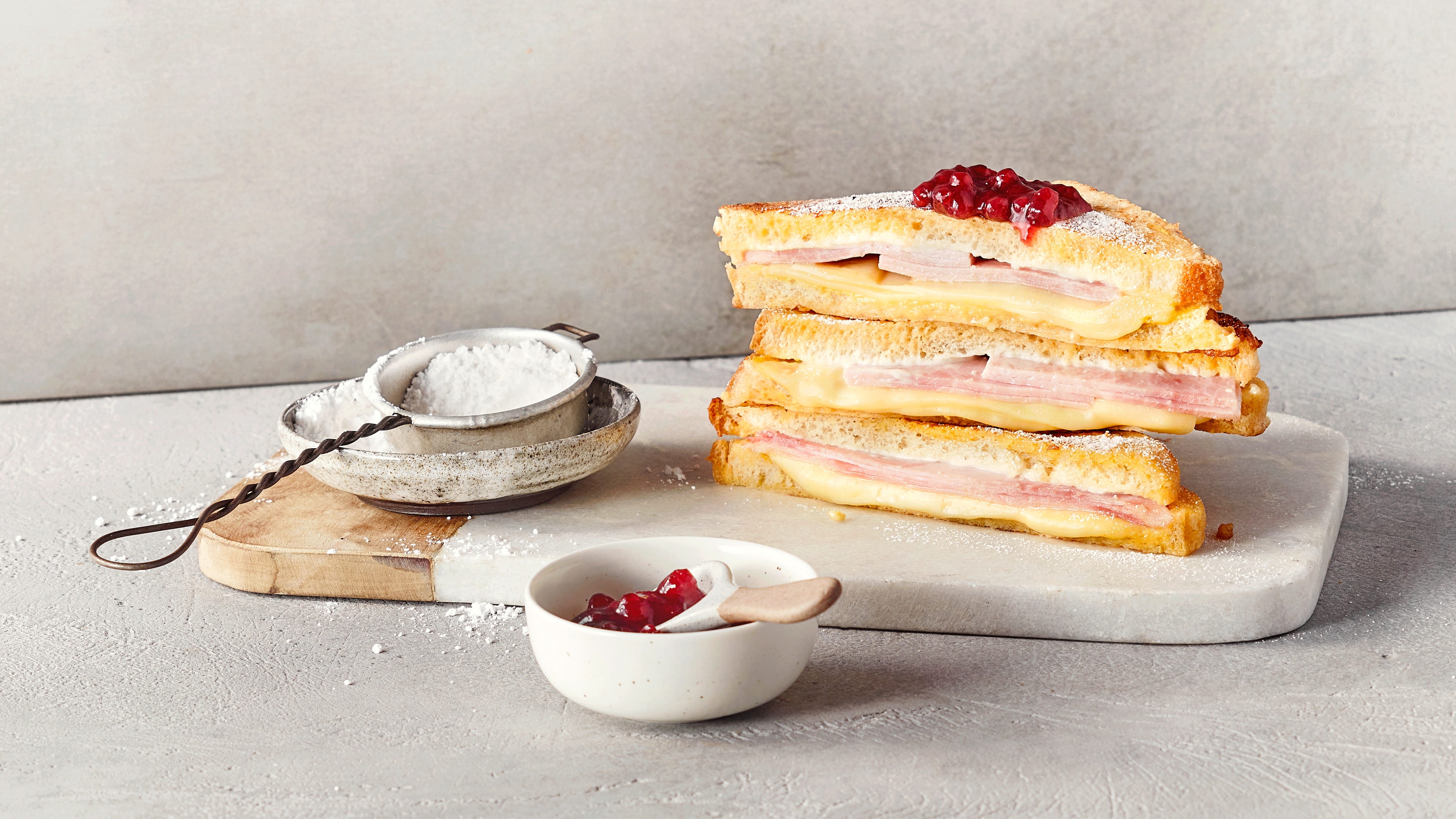 Ein Brett mit drei getoasteten Sandwiches, gefüllt mit Schinken und geschmolzenem Käse, garniert mit rotem Gelee..