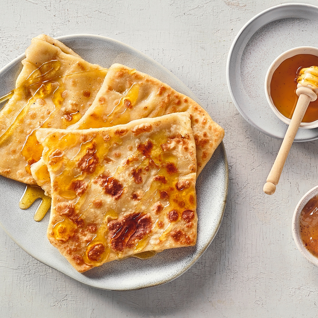 Ein Teller mit drei flachen, goldbraunen Teigstücken, die mit glänzendem Honig überzogen sind.