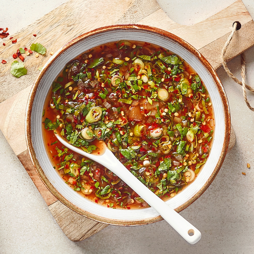 Eine Schüssel mit Brühe, darin grüne Kräuterstücke, grüne Zwiebelringe und kleinen roten Stückchen sowie einem weißen Löffel.