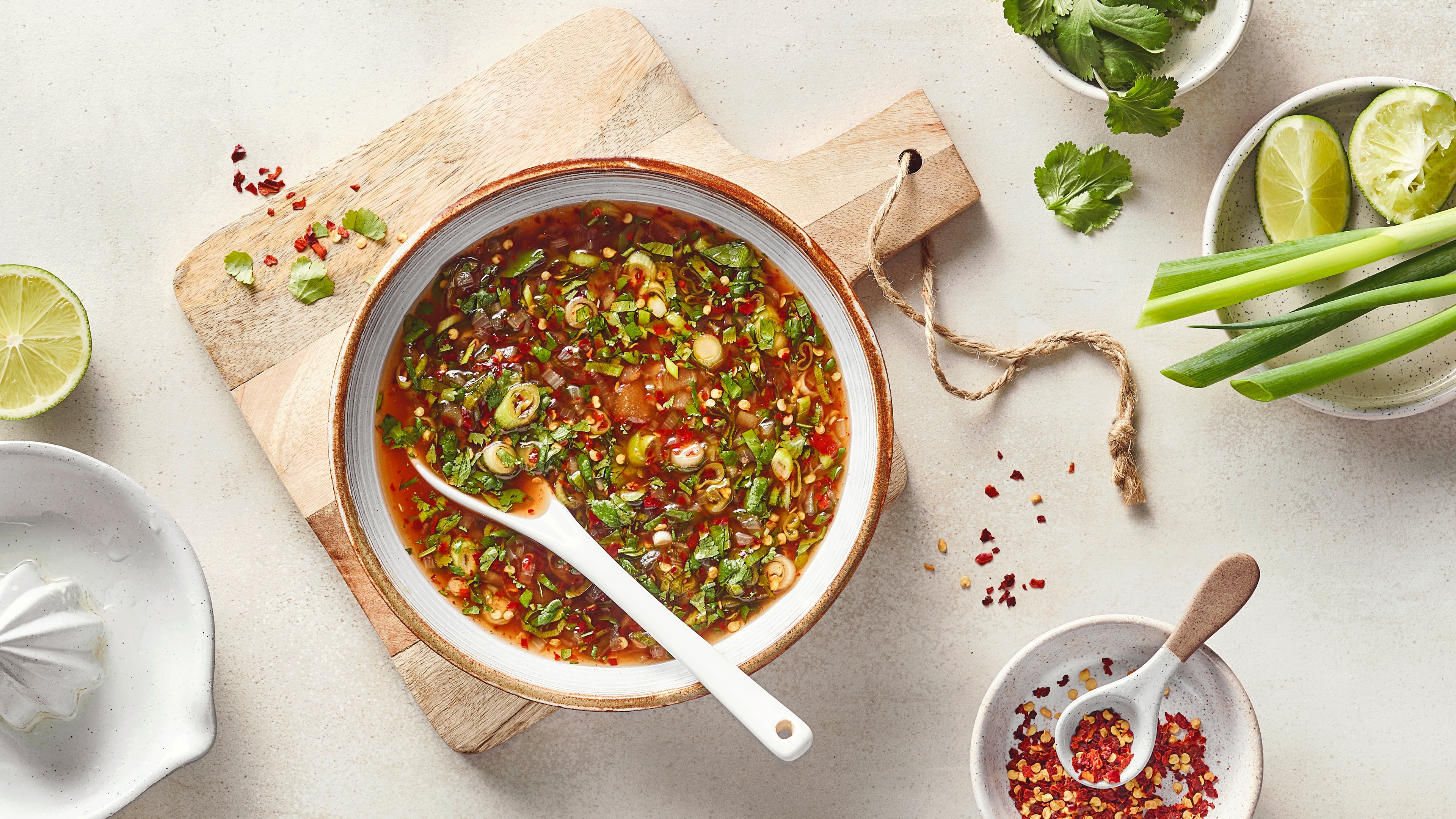 Eine Schüssel mit Brühe, darin grüne Kräuterstücke, grüne Zwiebelringe und kleinen roten Stückchen sowie einem weißen Löffel.