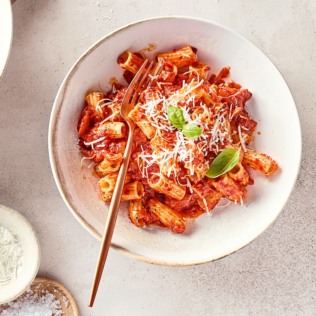 Ein Teller Rigatoni in Tomatensoße mit geriebenem Käse und Basilikum, serviert auf hellem Untergrund.