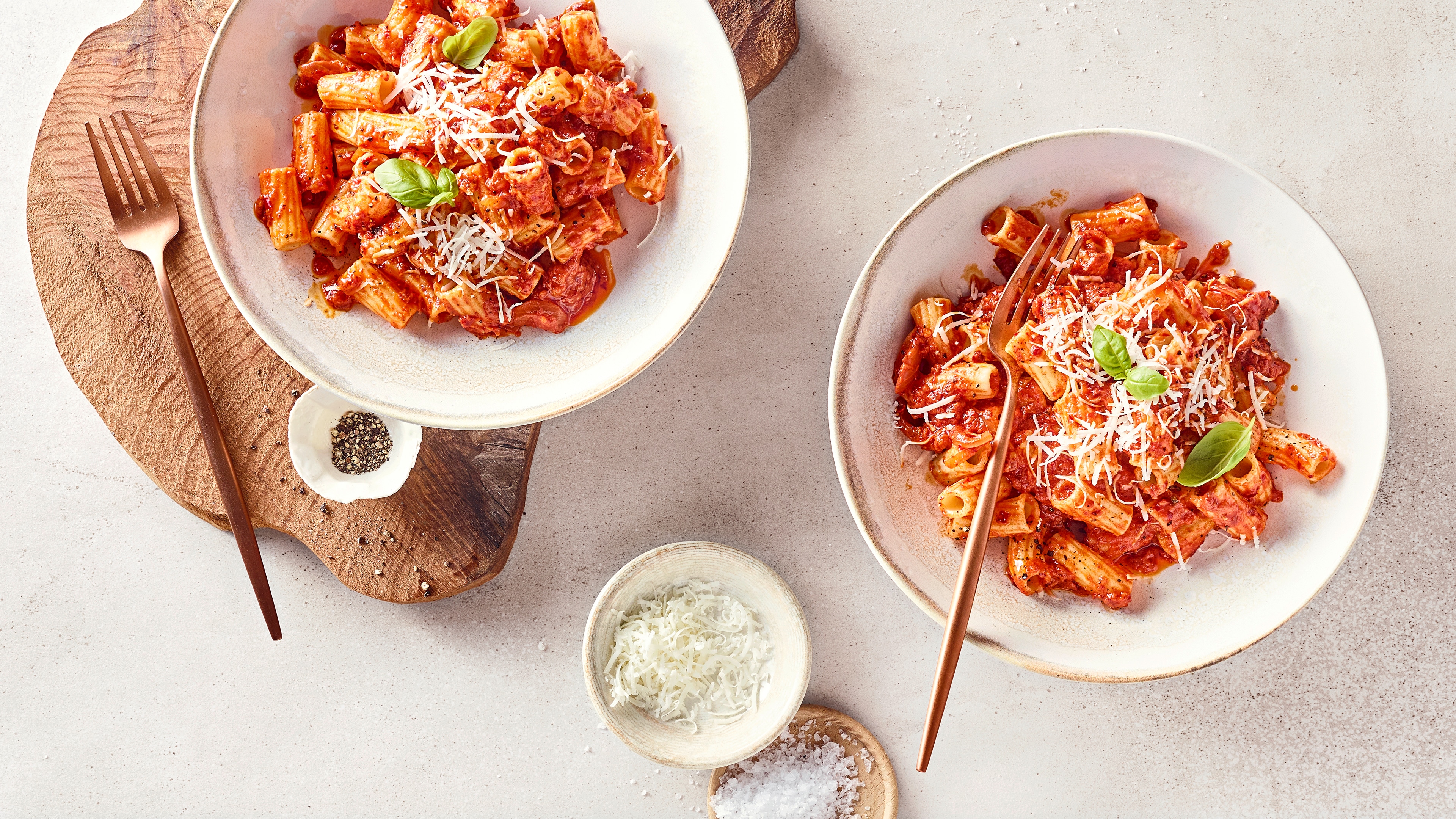 Zwei Teller Rigatoni in Tomatensoße mit geriebenem Käse und Basilikum, serviert auf hellem Untergrund.