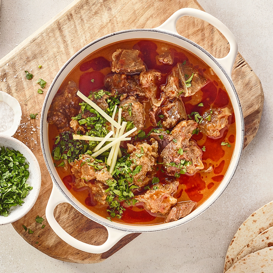 Ein Topf mit Fleischstücken in roter Soße, darauf grüne Kräuter & helle Streifen, daneben Schalen mit Salz und Kräutern.