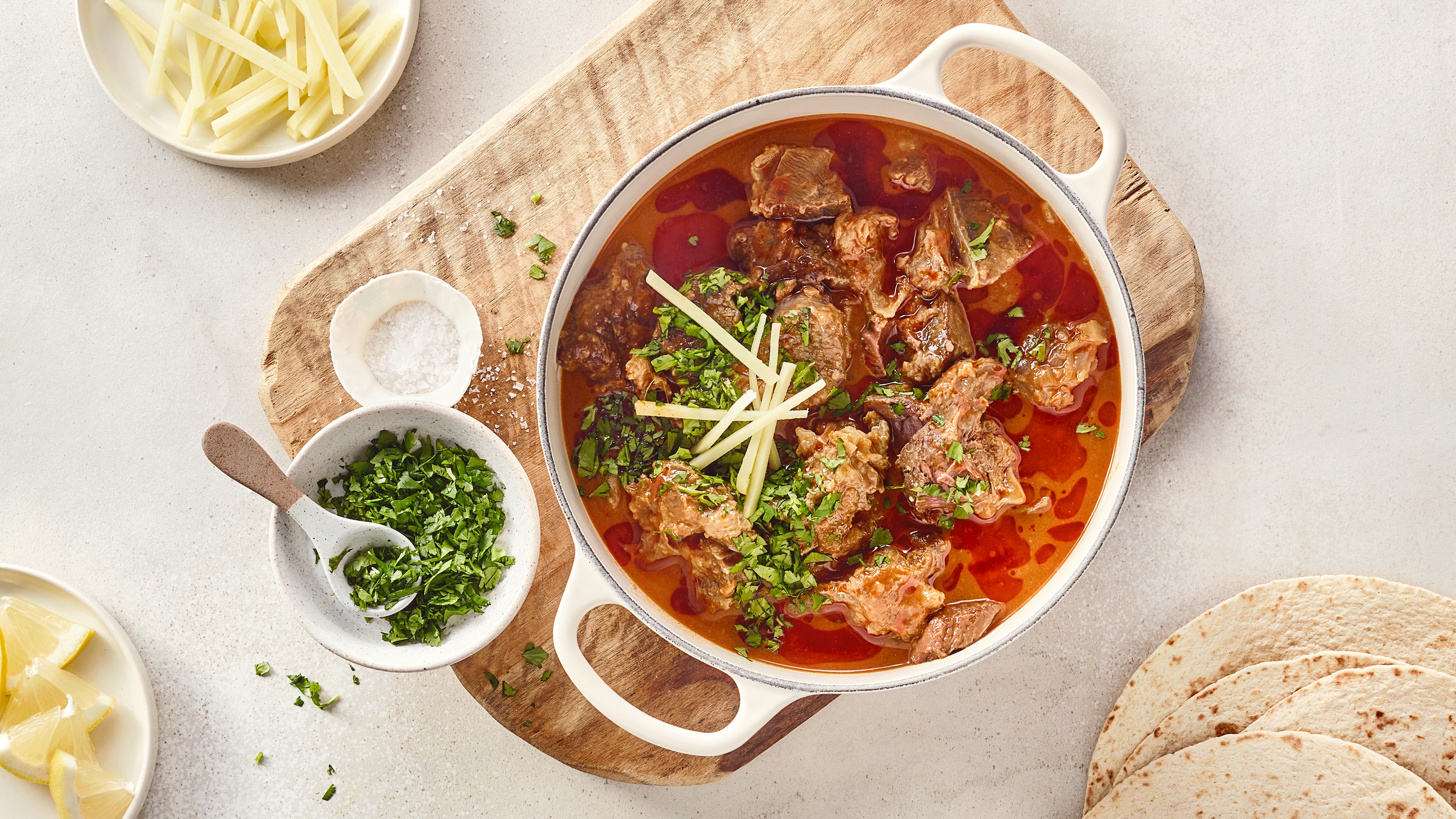 Ein Topf mit Fleischstücken in roter Soße, darauf grüne Kräuter & helle Streifen, daneben Schalen mit Salz und Kräutern.