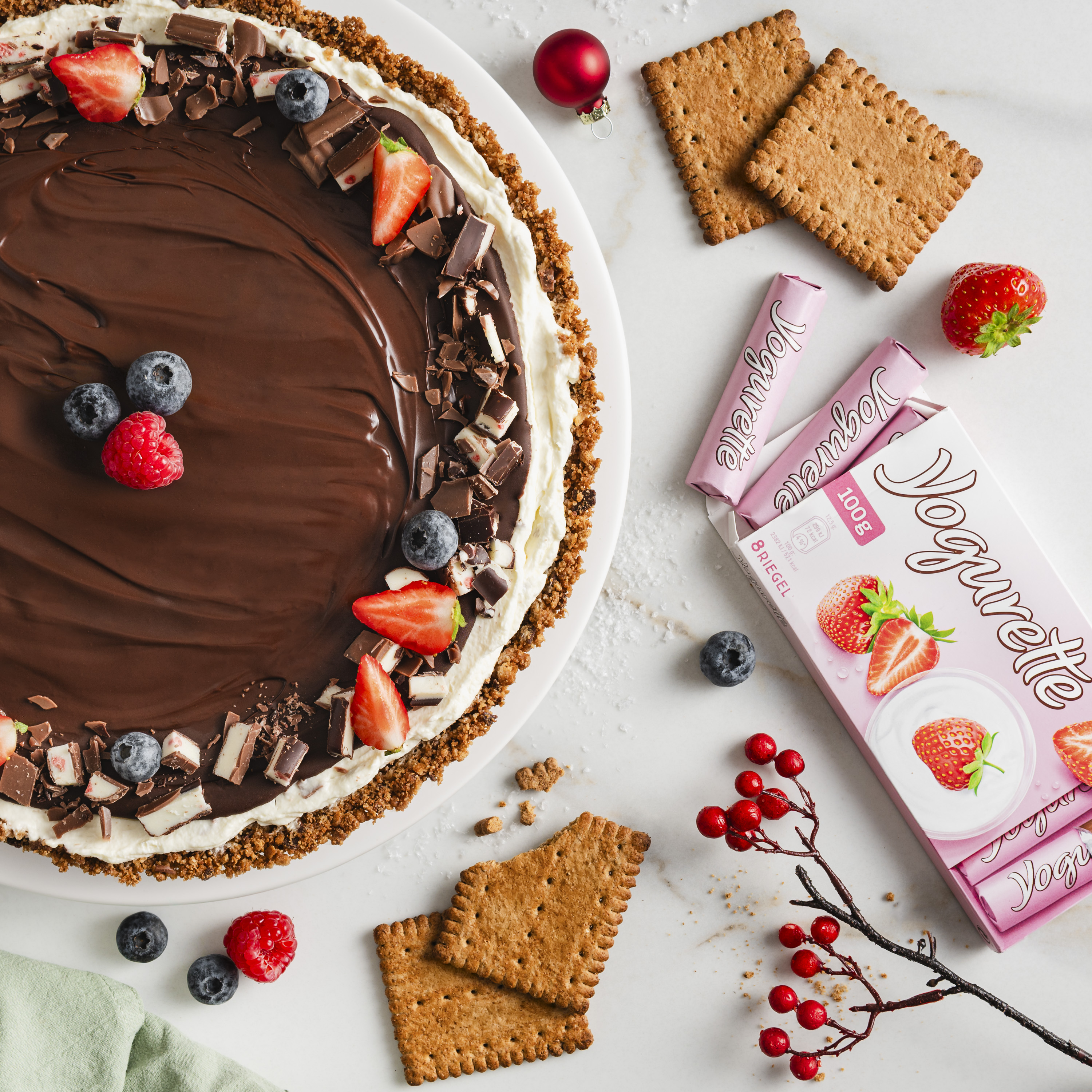 Ein Teller mit einer runden Torte auf krümeligem Keksboden, bedeckt mit glatter Schokoschicht, umrandet von Creme, Beeren und Schokostückchen
