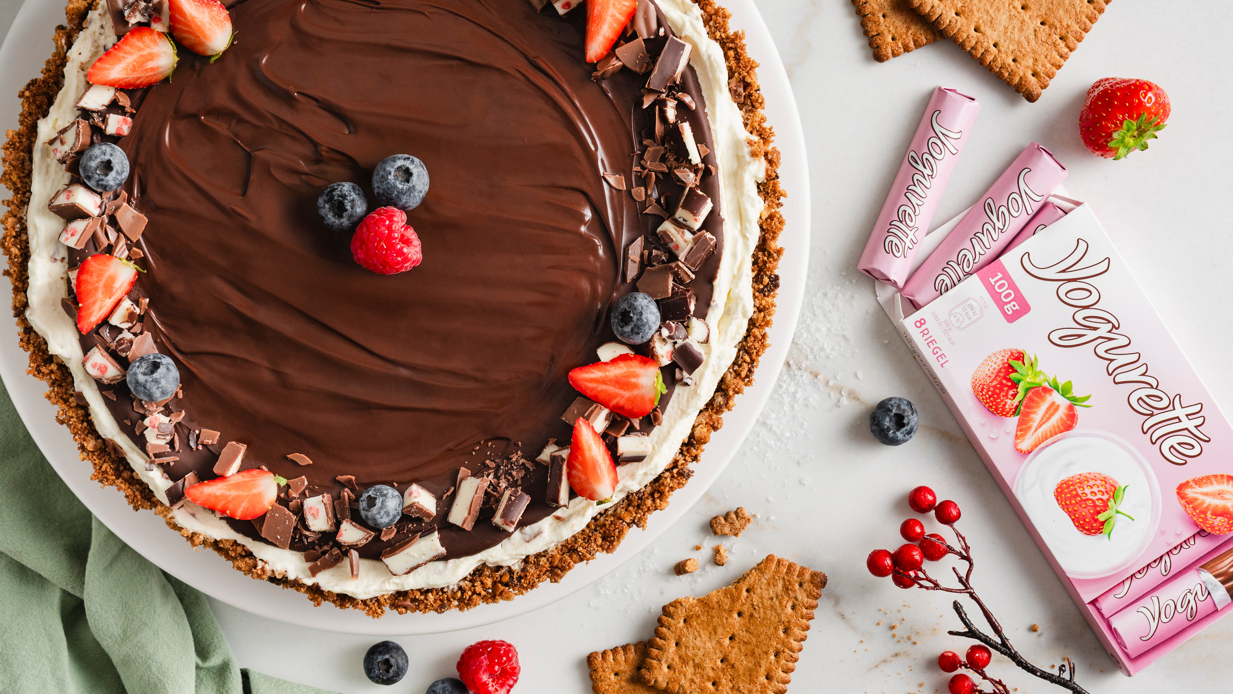 Ein Teller mit einer runden Torte auf krümeligem Keksboden, bedeckt mit glatter Schokoschicht, umrandet von Creme, Beeren und Schokostückchen