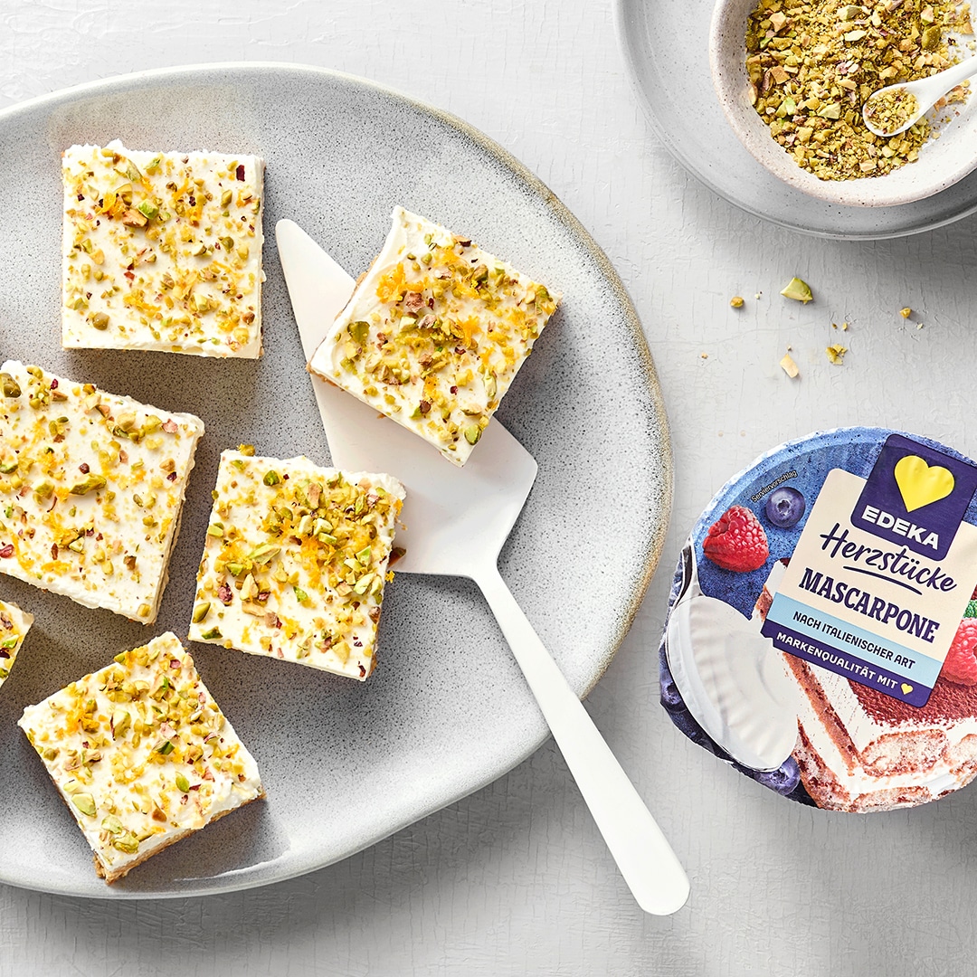 Mehrere viereckige Kuchenstücke mit Frosting und Pistazienstücken.  Oben rechts eine Schale mit gehackten Pistazien. Daneben das Produkt EDEKA Herzstücke Mascarpone