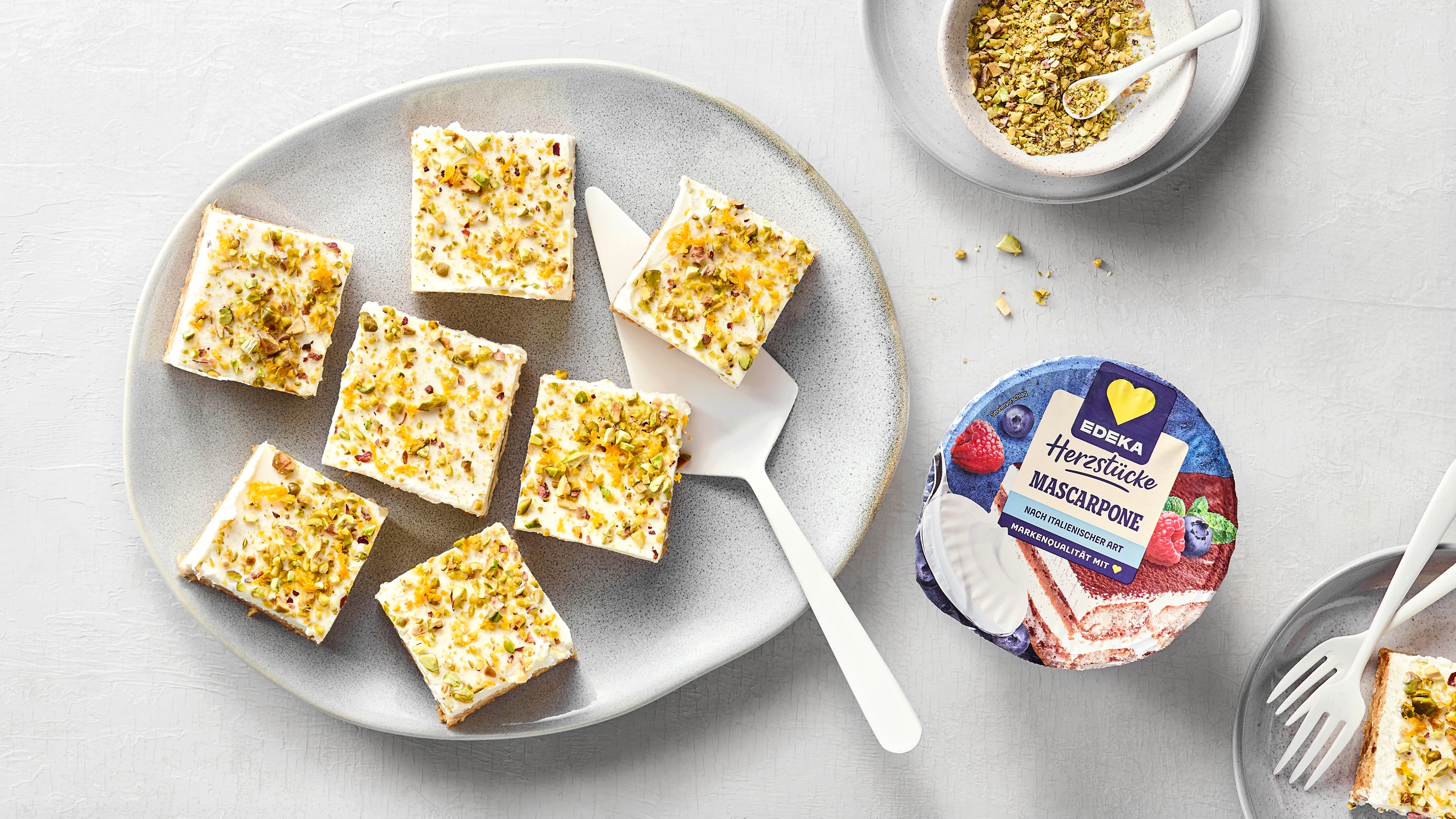 Mehrere viereckige Kuchenstücke mit Frosting und Pistazienstücken.  Oben rechts eine Schale mit gehackten Pistazien. Daneben das Produkt EDEKA Herzstücke Mascarpone