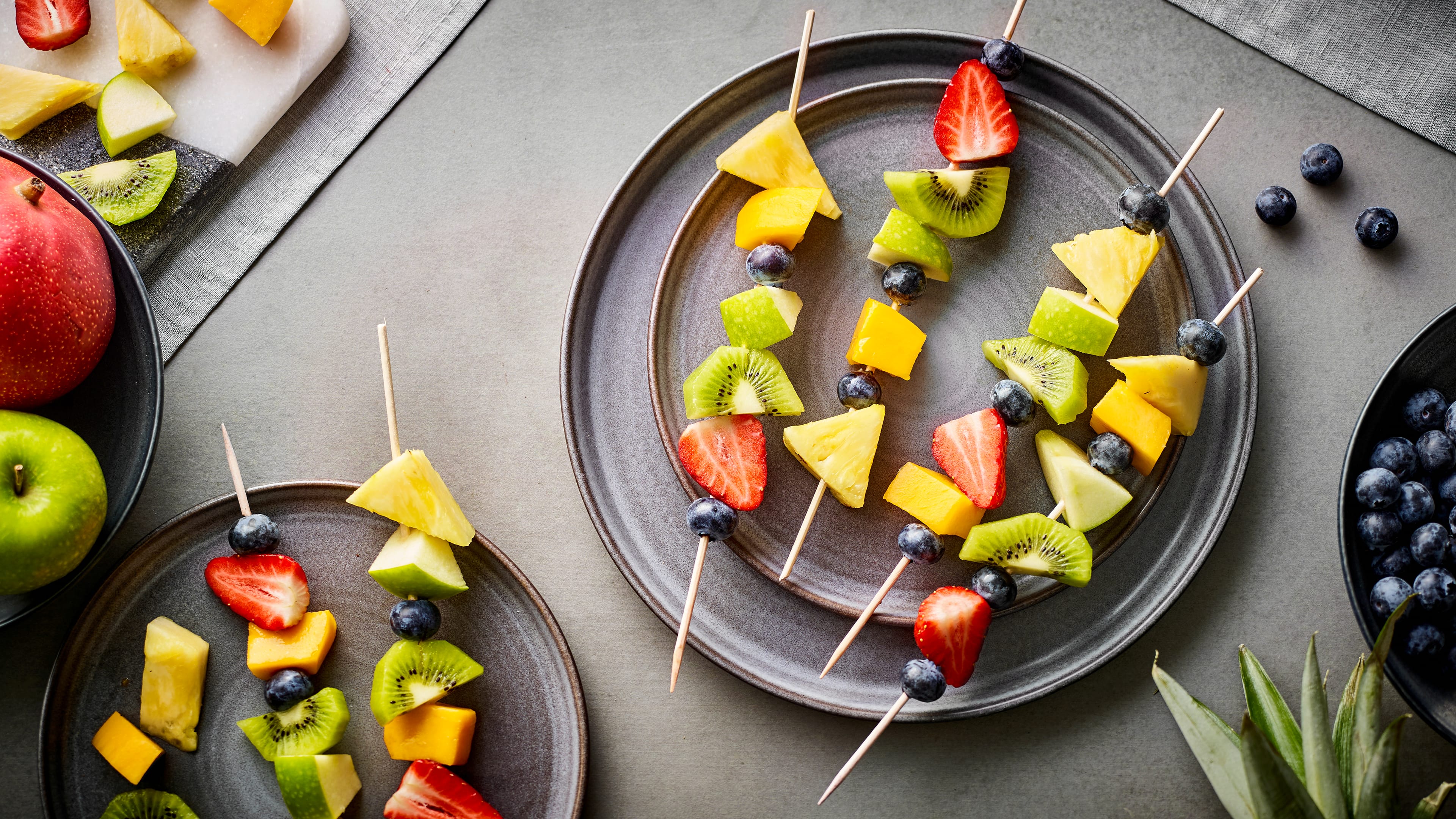 Rezept für Obstspieße Holzspieße mit Blaubeeren, Erdbeer-, Kiwi-, Ananas- und Mangostückchen, auf runden Tellern