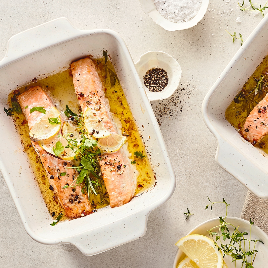 Kleine Auflaufformen mit gelbgold gebackenem Lachs in Öl, getoppt mit frischen Kräutern und Zitronenspalten