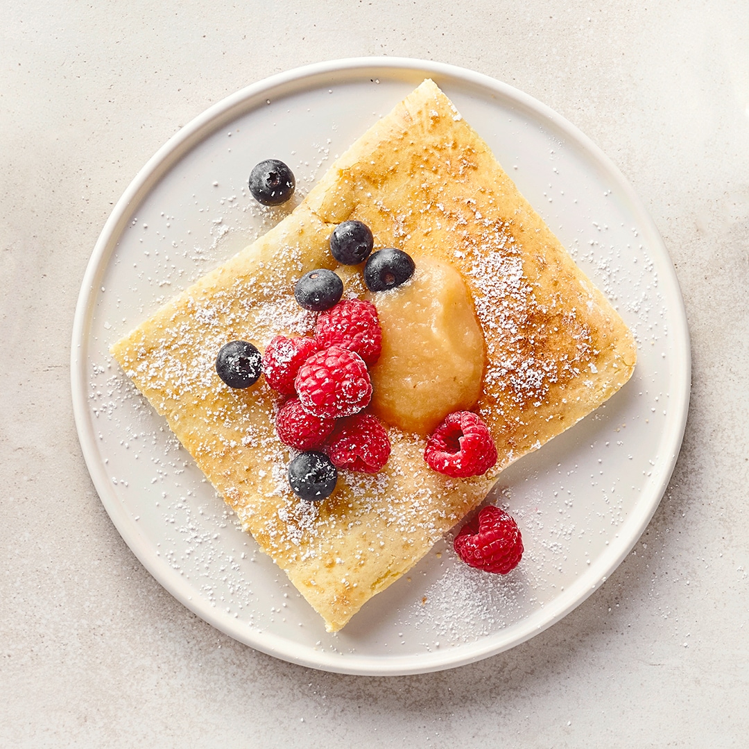 Ein Teller mit einem Stück Pfannkuchen, bestreut mit Puderzucker, Blaubeeren, Himbeeren und einem Klecks Apfelmus
