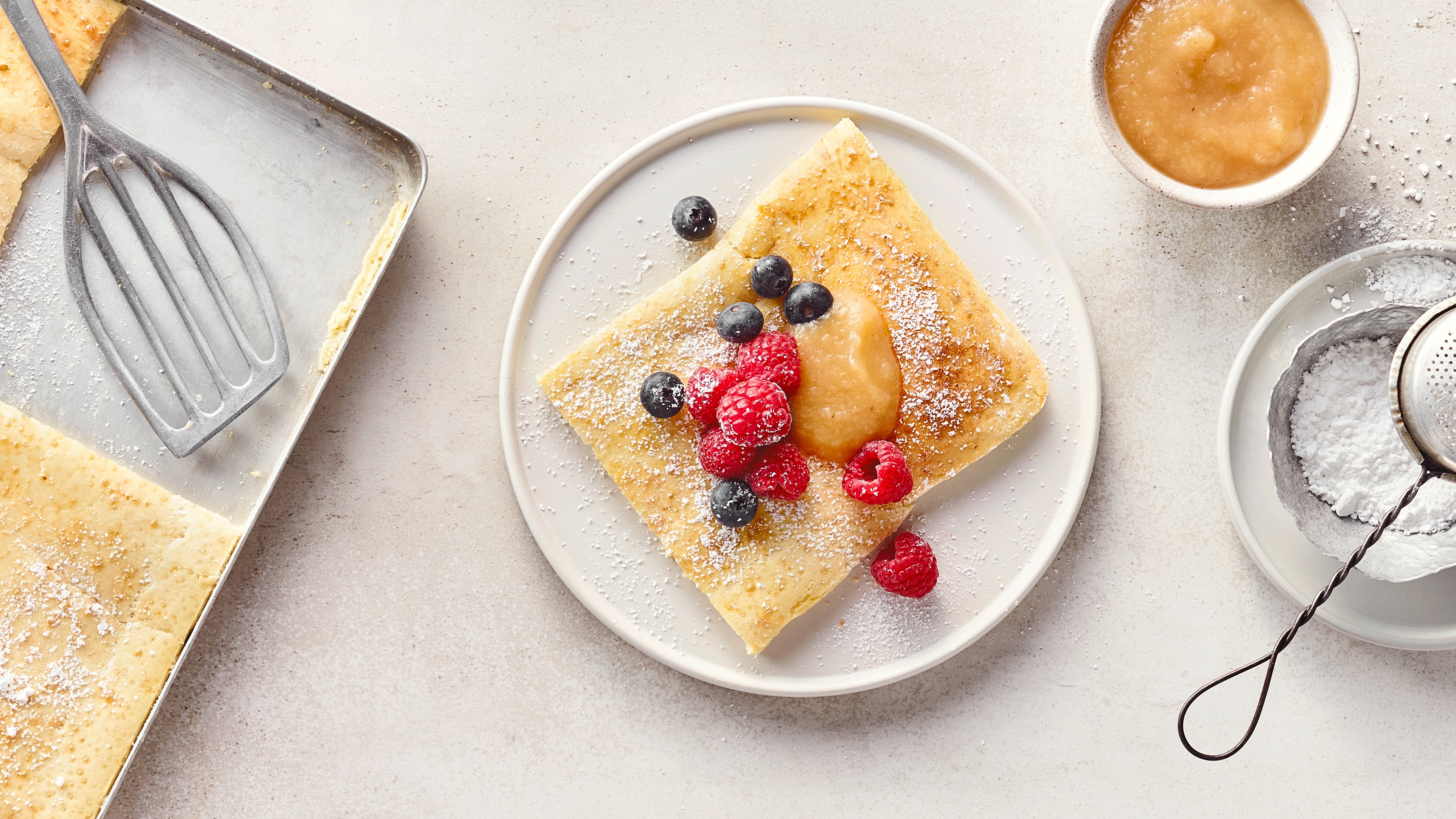 Rezept für Ofenpfannkuchen Ein Teller mit einem Stück Pfannkuchen, bestreut mit Puderzucker, Blaubeeren, Himbeeren und einem Klecks Apfelmus