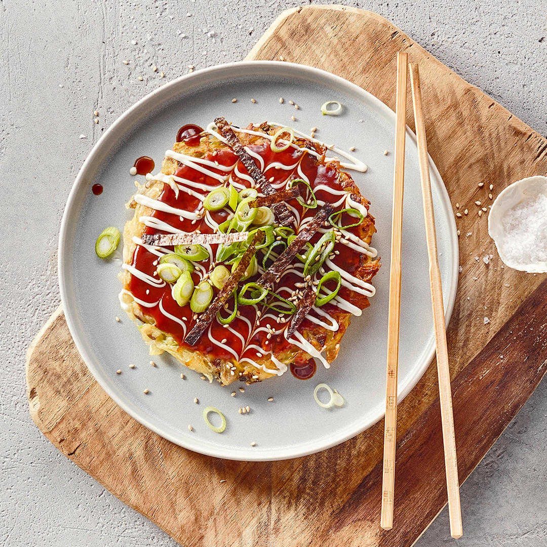Ein Teller mit einem runden Pfannkuchen, bedeckt mit braunen Streifen, grünen Lauchringen, roter und weißer Soße sowie Sesam.
