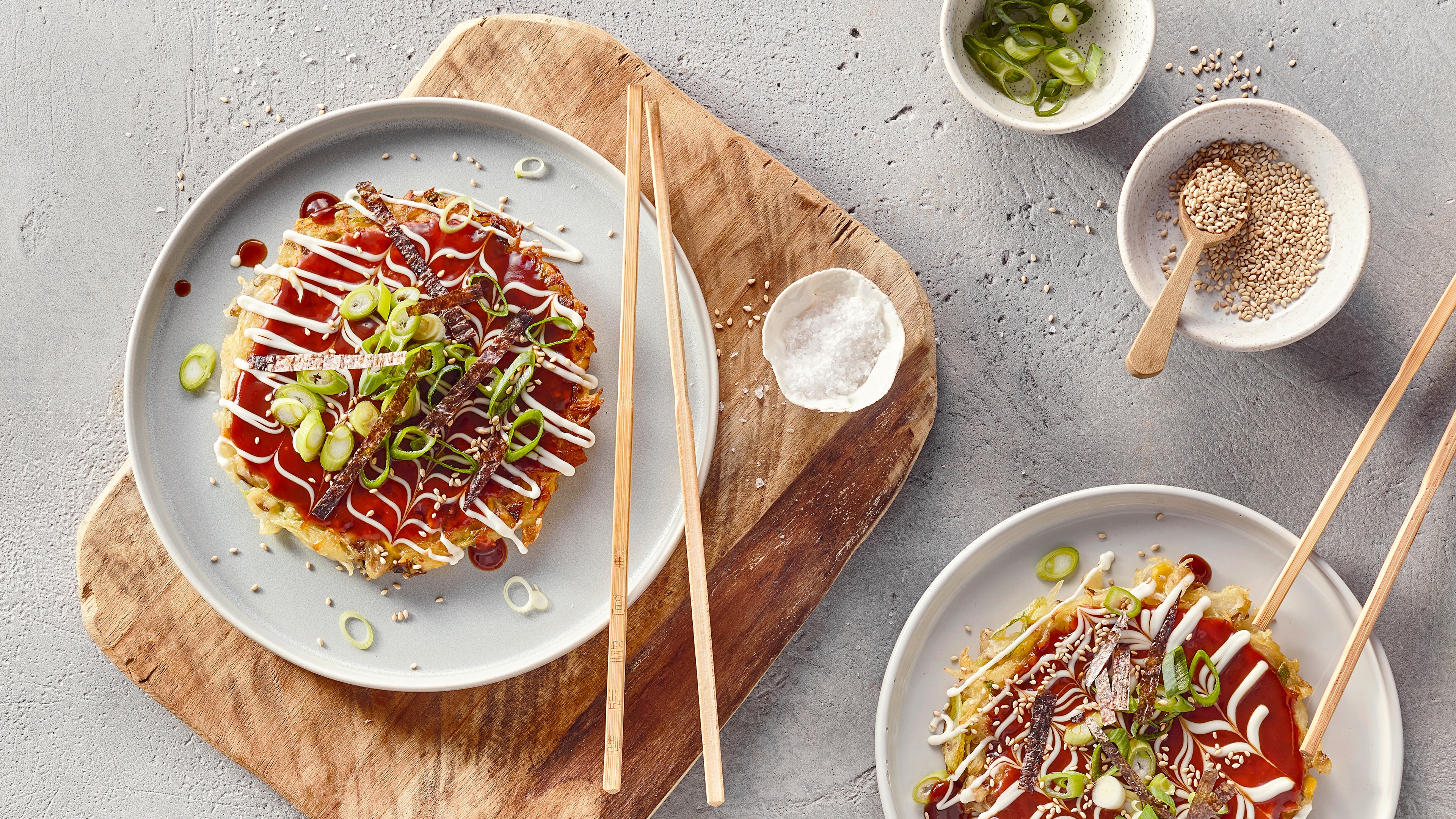 Ein Teller mit einem runden Pfannkuchen, bedeckt mit braunen Streifen, grünen Lauchringen, roter und weißer Soße sowie Sesam.