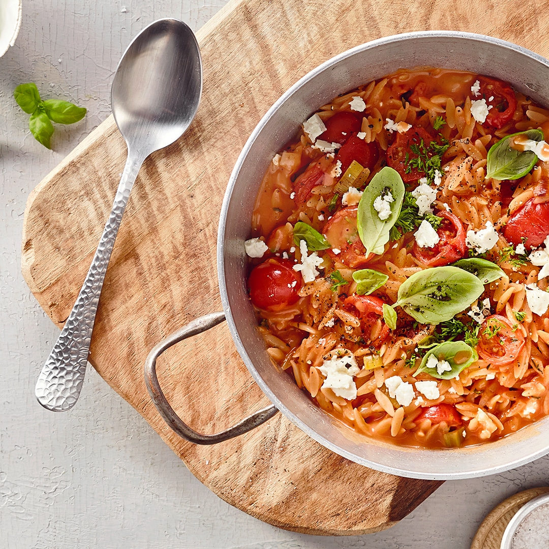 Ein Topf mit kleinen Nudeln in roter Soße, darin rote Tomatenhälften, grüne Basilikumblätter und weiße Käsekrümel.