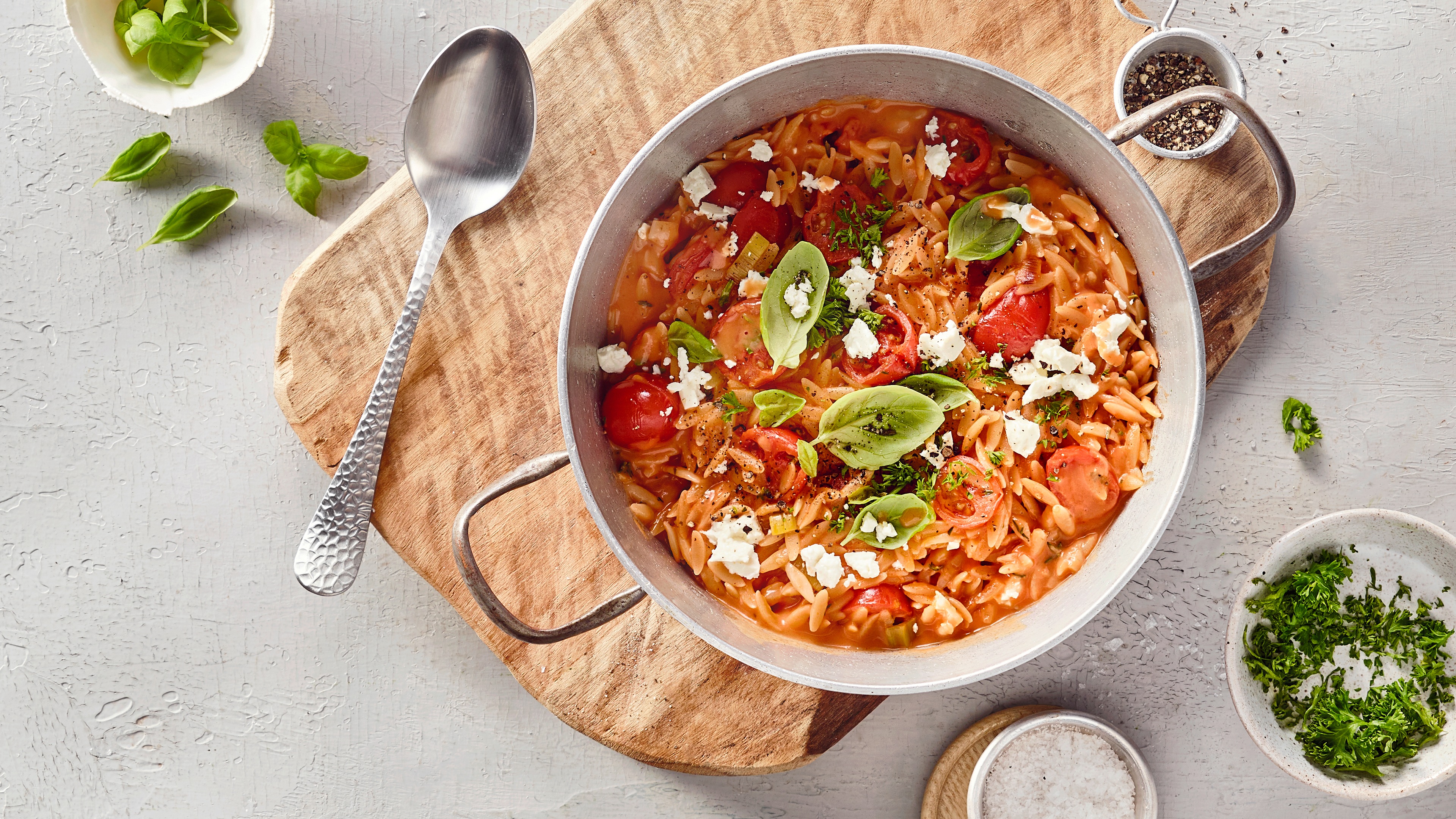 Ein Topf mit kleinen Nudeln in roter Soße, darin rote Tomatenhälften, grüne Basilikumblätter und weiße Käsekrümel.