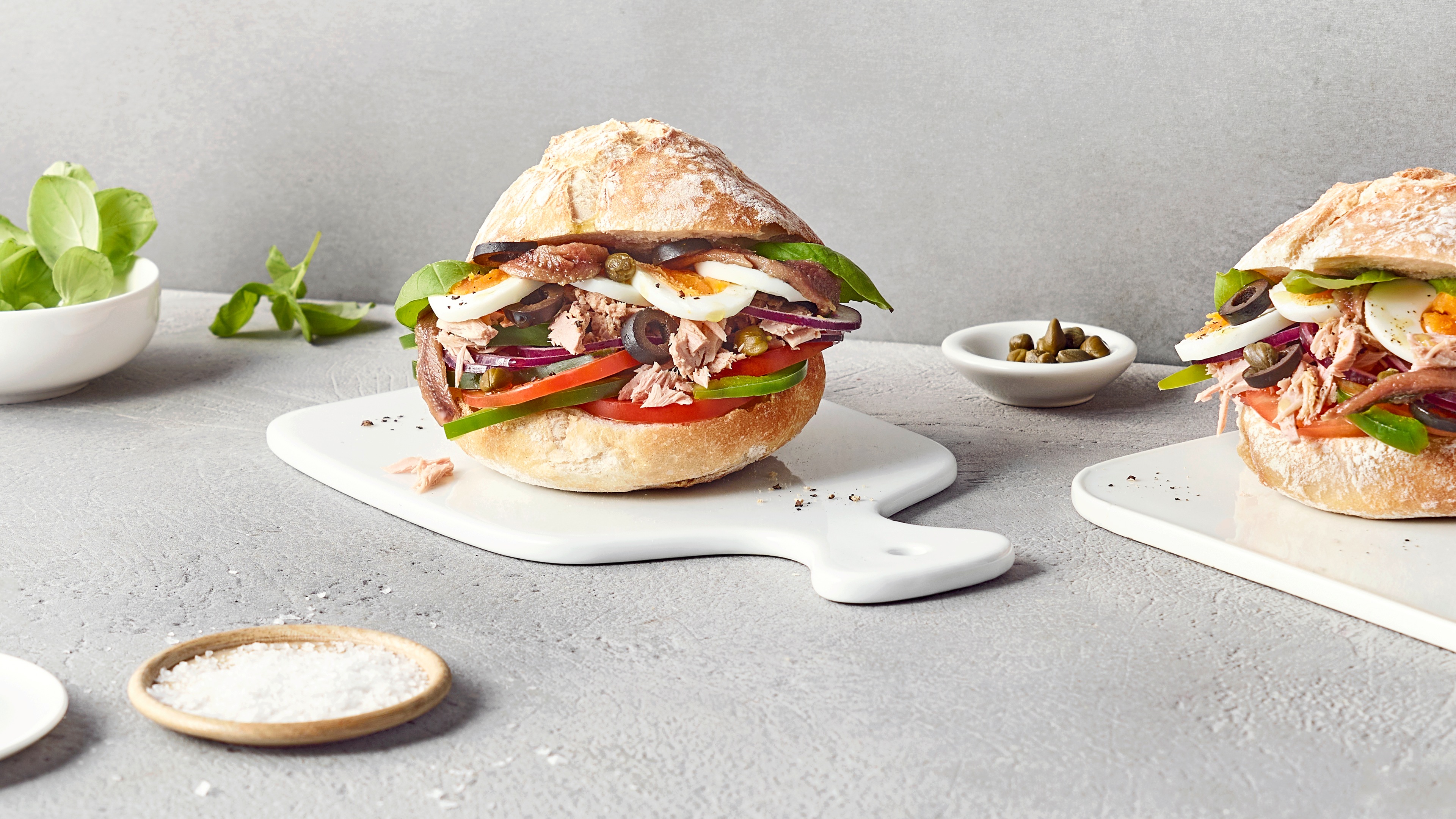 Ein Teller mit einem Brötchen, gefüllt mit Thunfisch, gekochten Eierscheiben, Tomaten, grünen Paprikastreifen, roten Zwiebeln, Kapern und Sardellen.