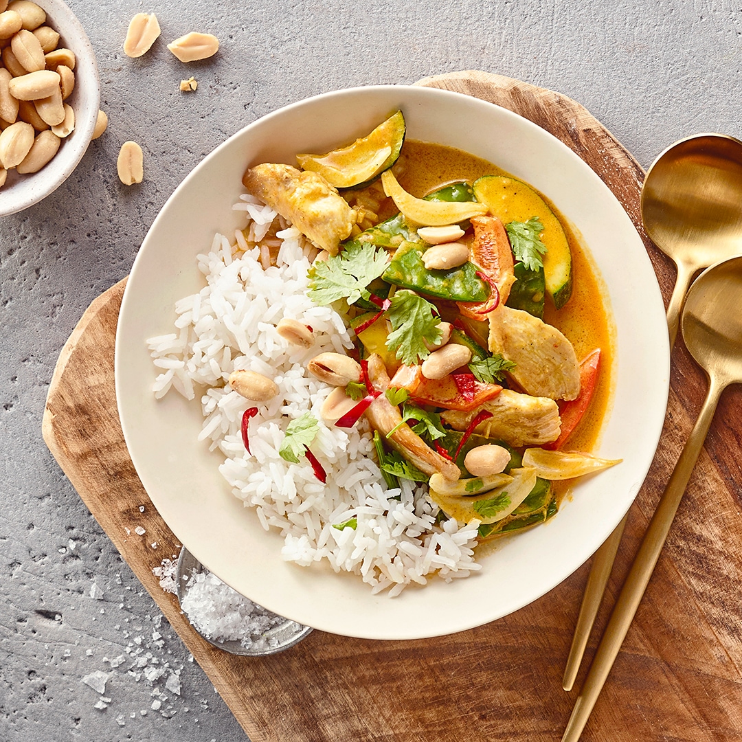 Eine Schale mit weißem Reis und Curry aus Hähnchen, Paprika, Zucchini, Koriander und Erdnüssen, daneben zwei goldene Löffel