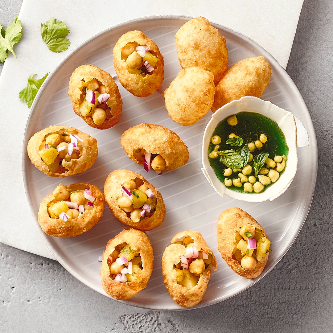 Ein Teller mit elf knusprigen Teigschalen, gefüllt mit Kichererbsen, Kartoffelstücken und Zwiebeln, daneben eine Schale mit grüner Sauce