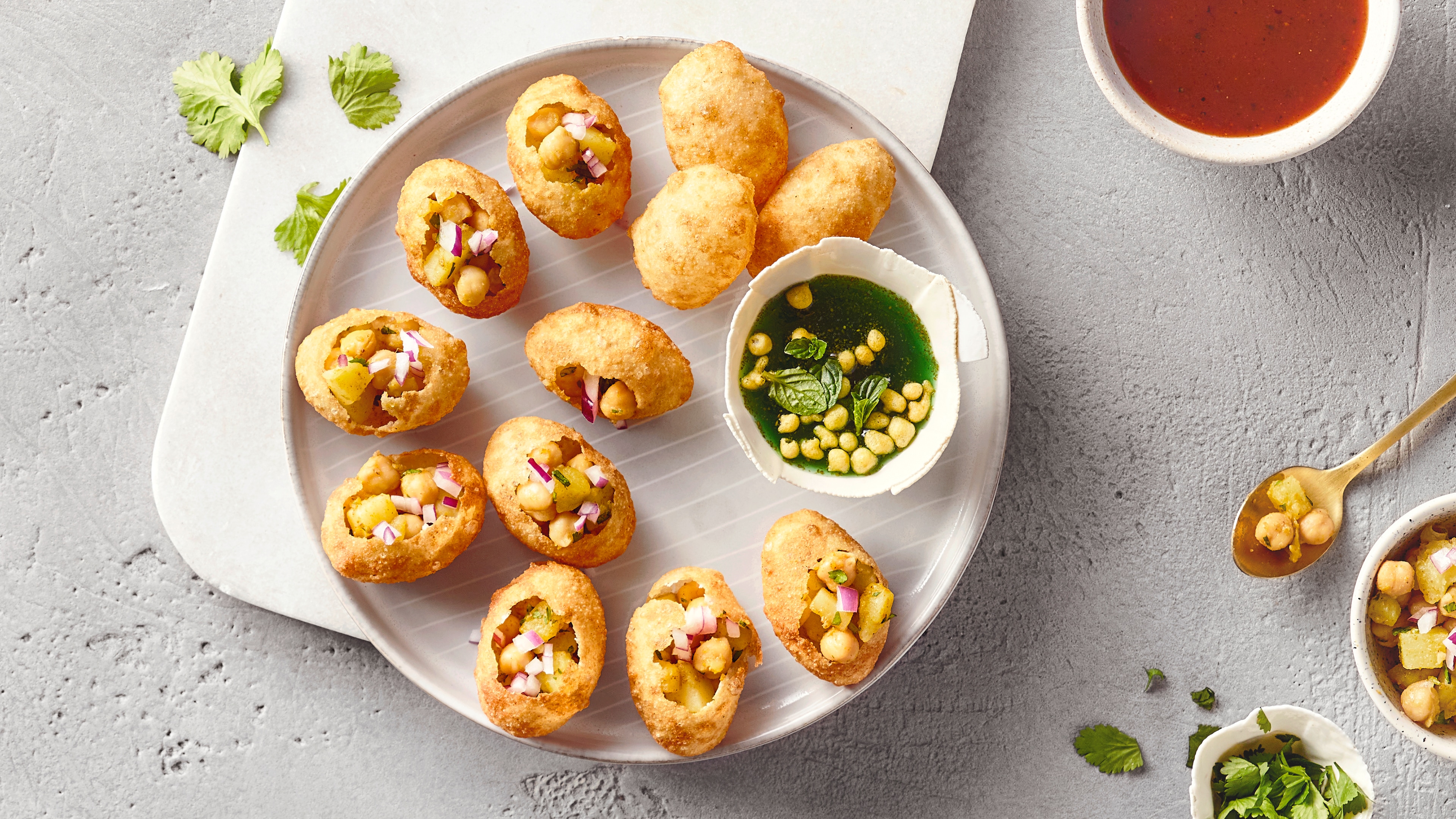 Ein Teller mit elf knusprigen Teigschalen, gefüllt mit Kichererbsen, Kartoffelstücken und Zwiebeln, daneben eine Schale mit grüner Sauce