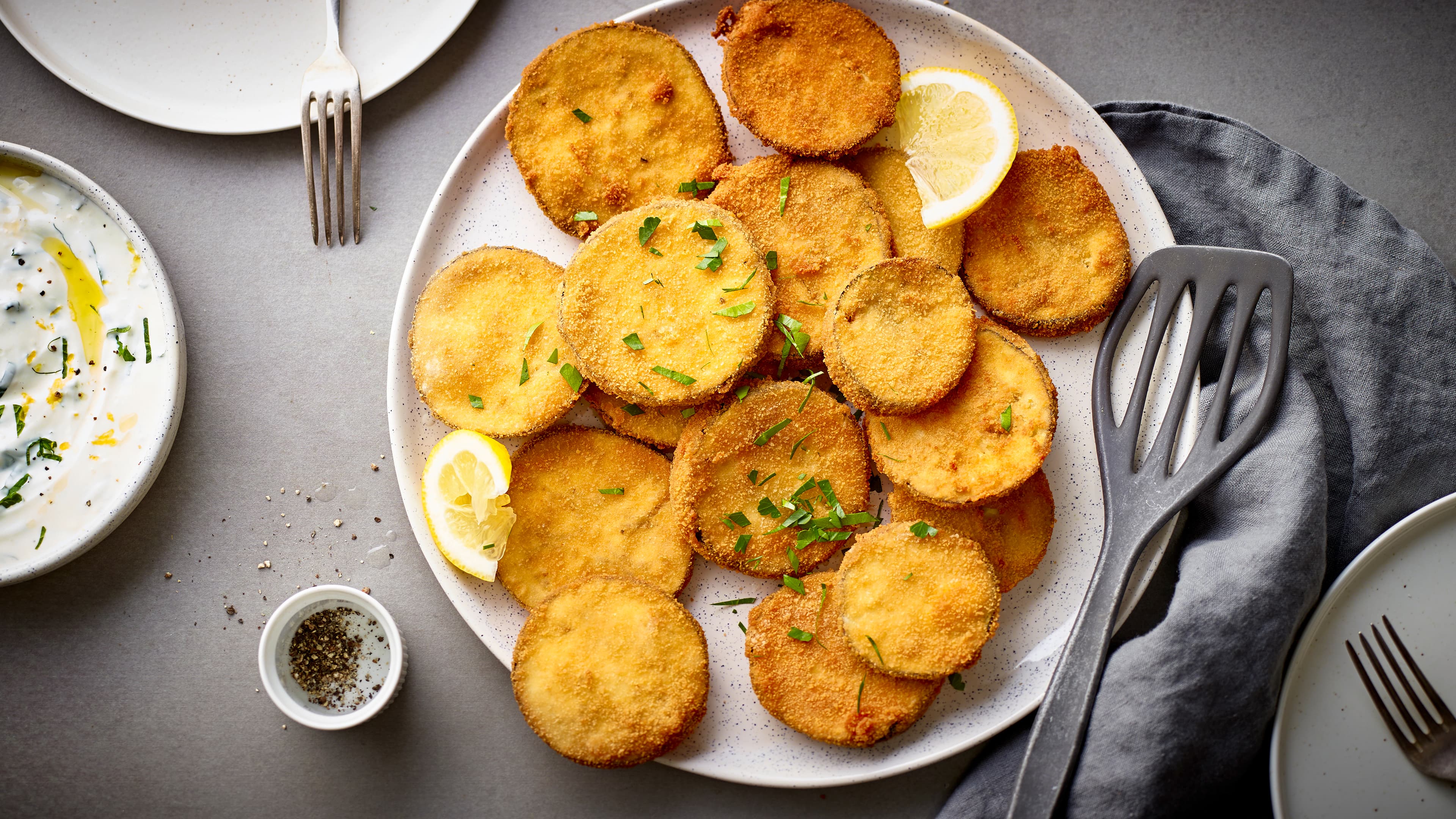 Rezept für panierte Aubergine Ein heller Teller, darauf in Panade frittierte runde Taler, garniert mit Kräutern und Zitronen, daneben ein Pfannenwender