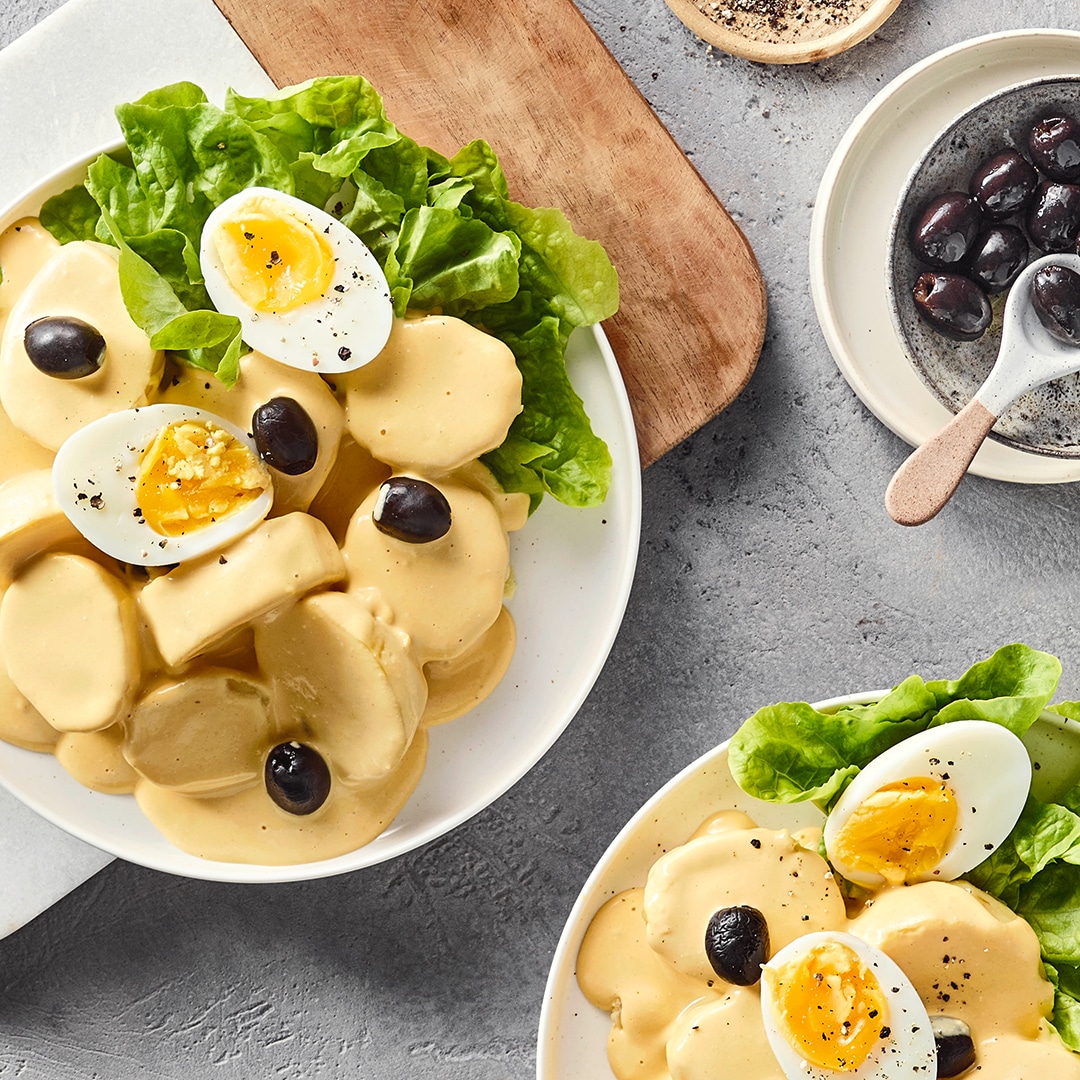 Ein Teller mit hellgelben Soßenscheiben, grünen Salatblättern, halbierten gekochten Eiern und schwarzen Oliven.