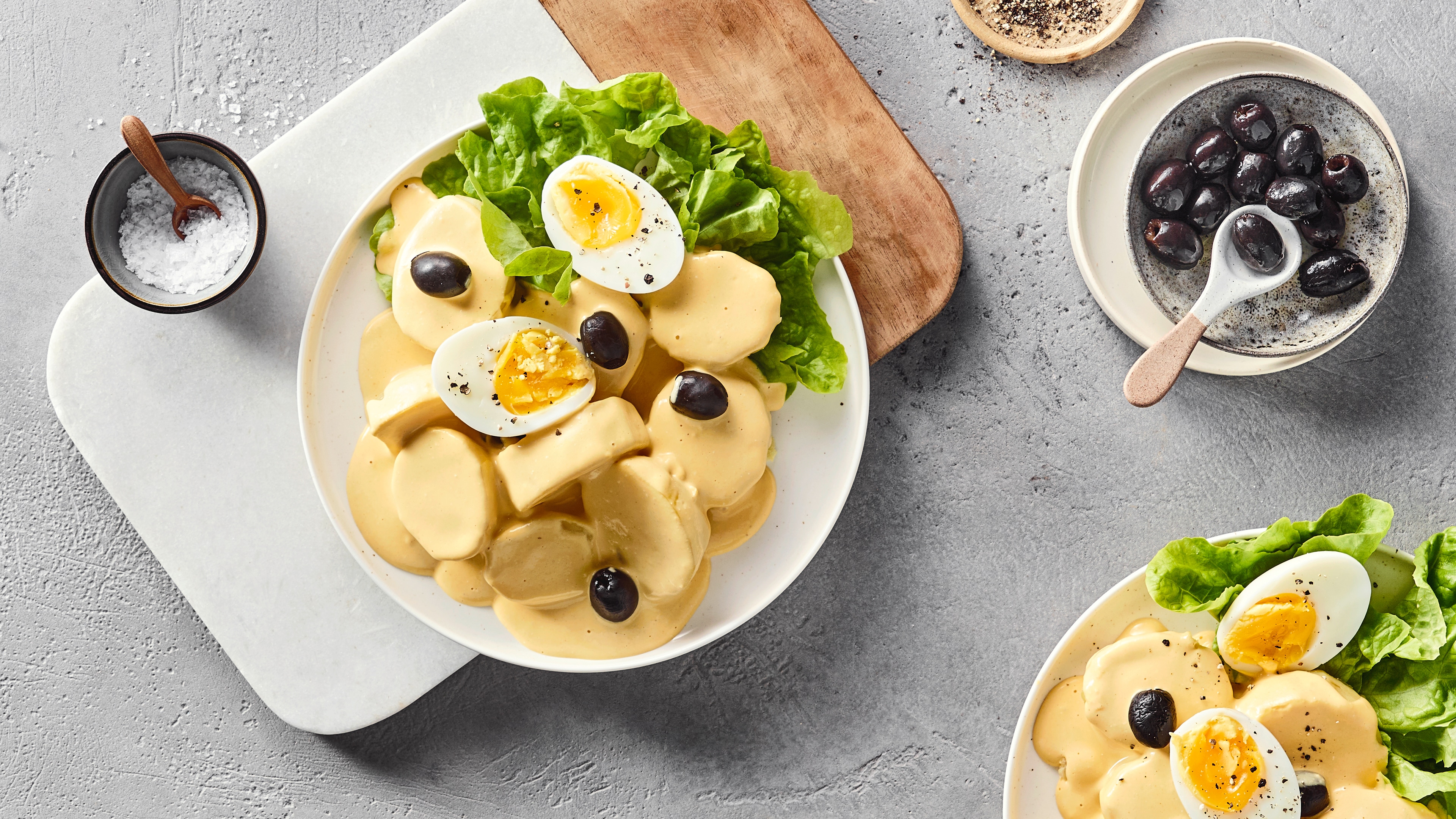 Ein Teller mit hellgelben Soßenscheiben, grünen Salatblättern, halbierten gekochten Eiern und schwarzen Oliven.