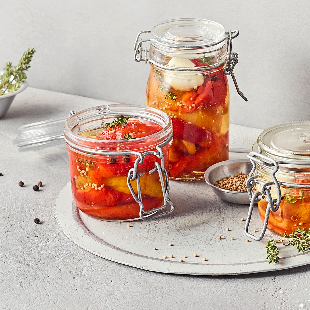 Ein Glas mit eingelegten roten und gelben Paprikastreifen in klarer Flüssigkeit, mit Gewürzkörnern und Thymian.