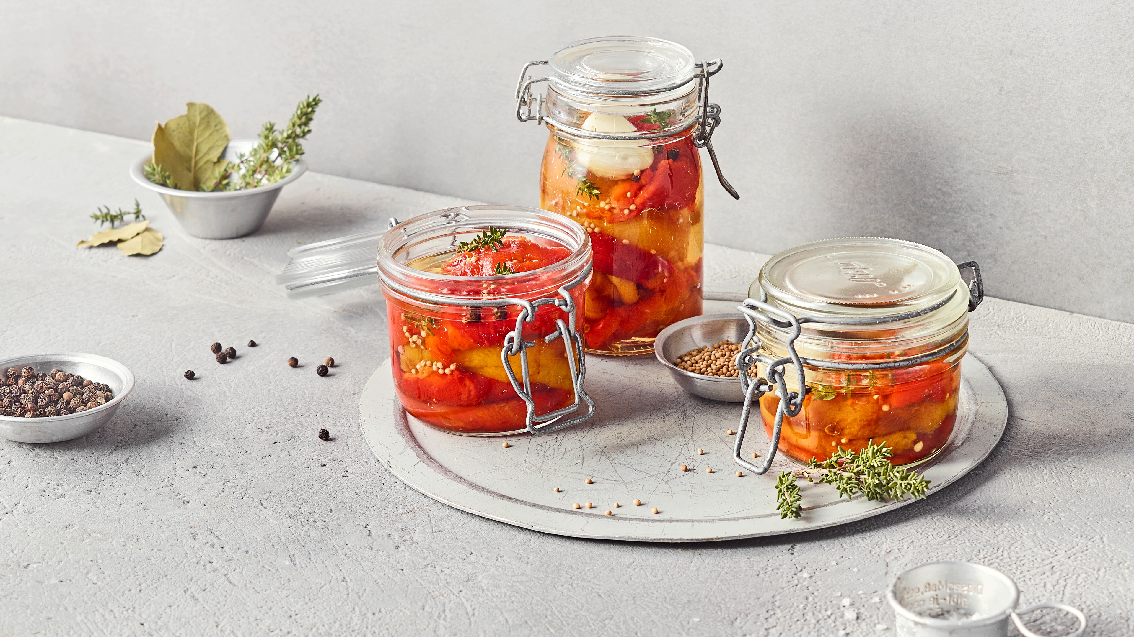 Ein Glas mit eingelegten roten und gelben Paprikastreifen in klarer Flüssigkeit, mit Gewürzkörnern und Thymian.