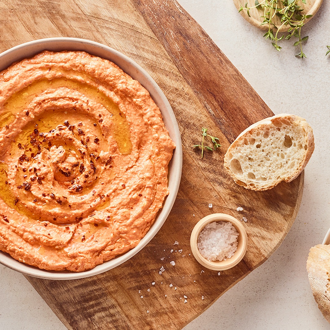Eine kleine Schüssel mit cremigem hellorangenem Dipp, getoppt mit etwas Öl, daneben ein paar Scheiben frisches Baguette