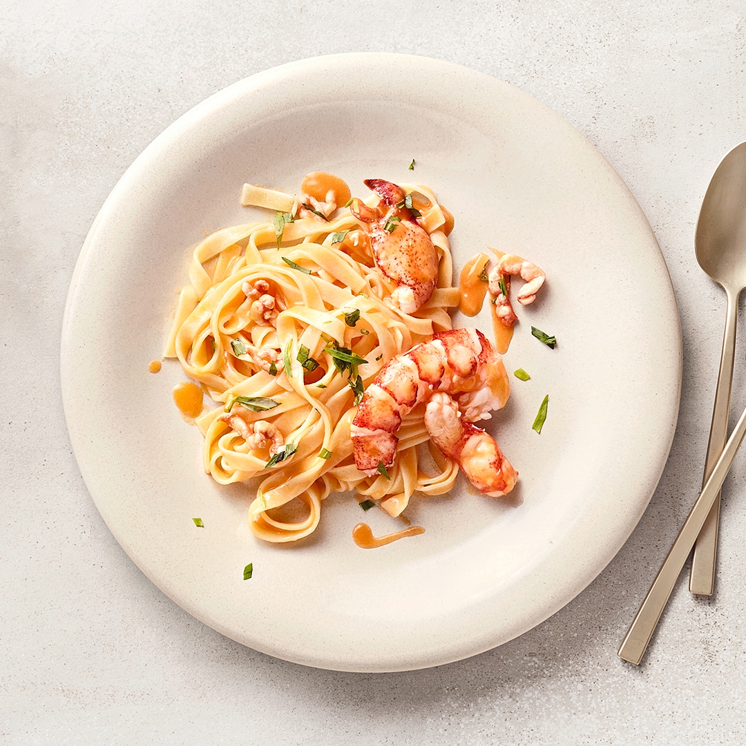 Tagliatelle mit Hummerstücken in cremiger Sauce, angerichtet auf einem weißen Teller und mit frischen Kräutern garniert.