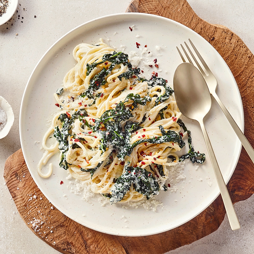 Ein Teller mit Spaghetti in heller Soße, darauf dunkle Blattstücke, geriebener Käse und rote Chiliflocken.