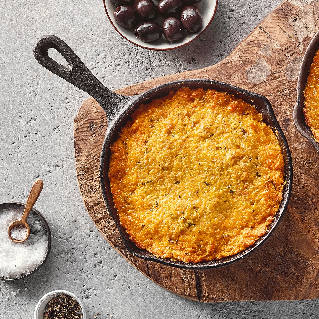 Eine schwarze Pfanne mit einer goldgelb überbackenen Masse, leicht gebräunten Rändern, daneben eine Schale mit Oliven.