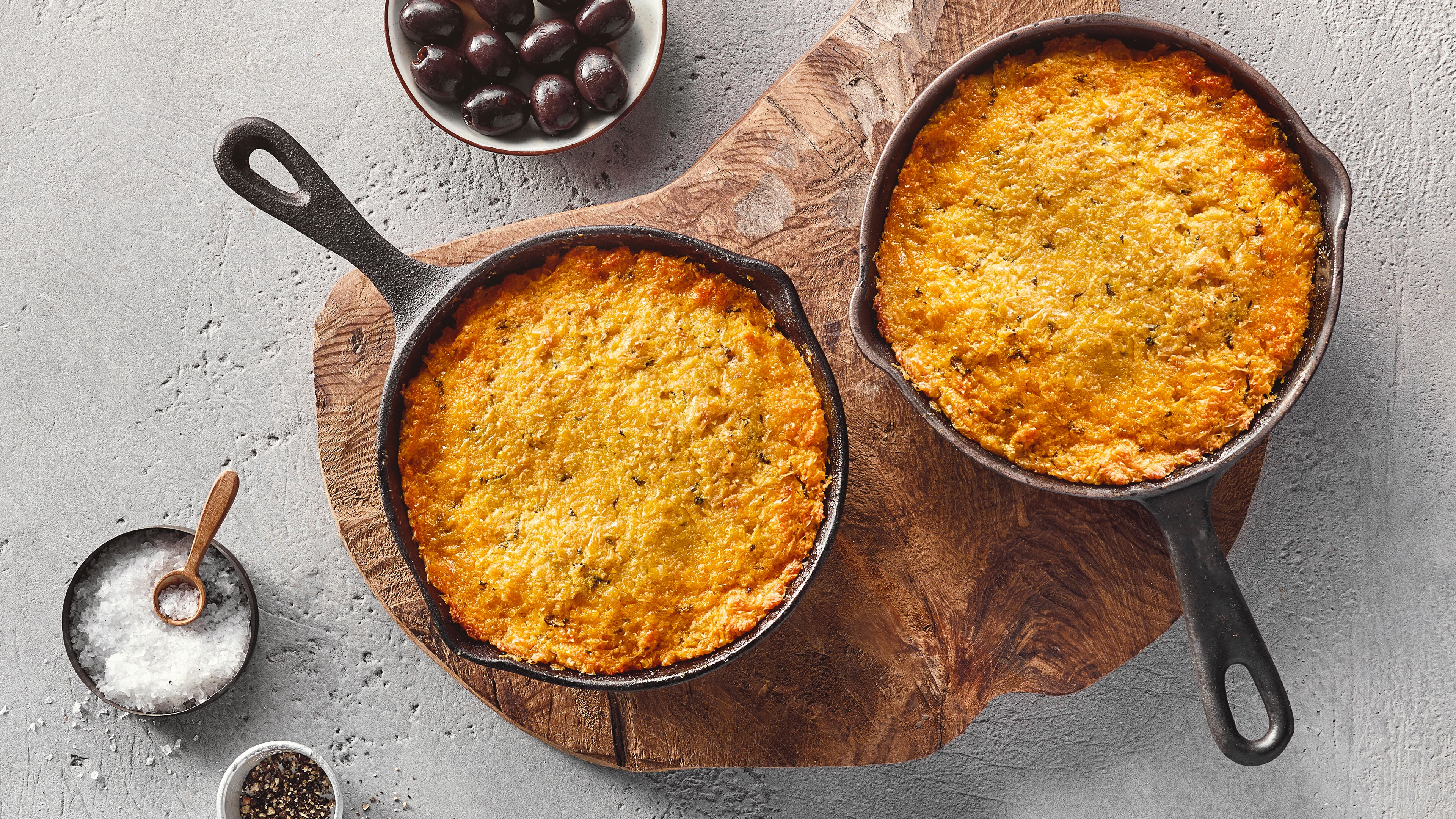 Eine schwarze Pfanne mit einer goldgelb überbackenen Masse, leicht gebräunten Rändern, daneben eine Schale mit Oliven.