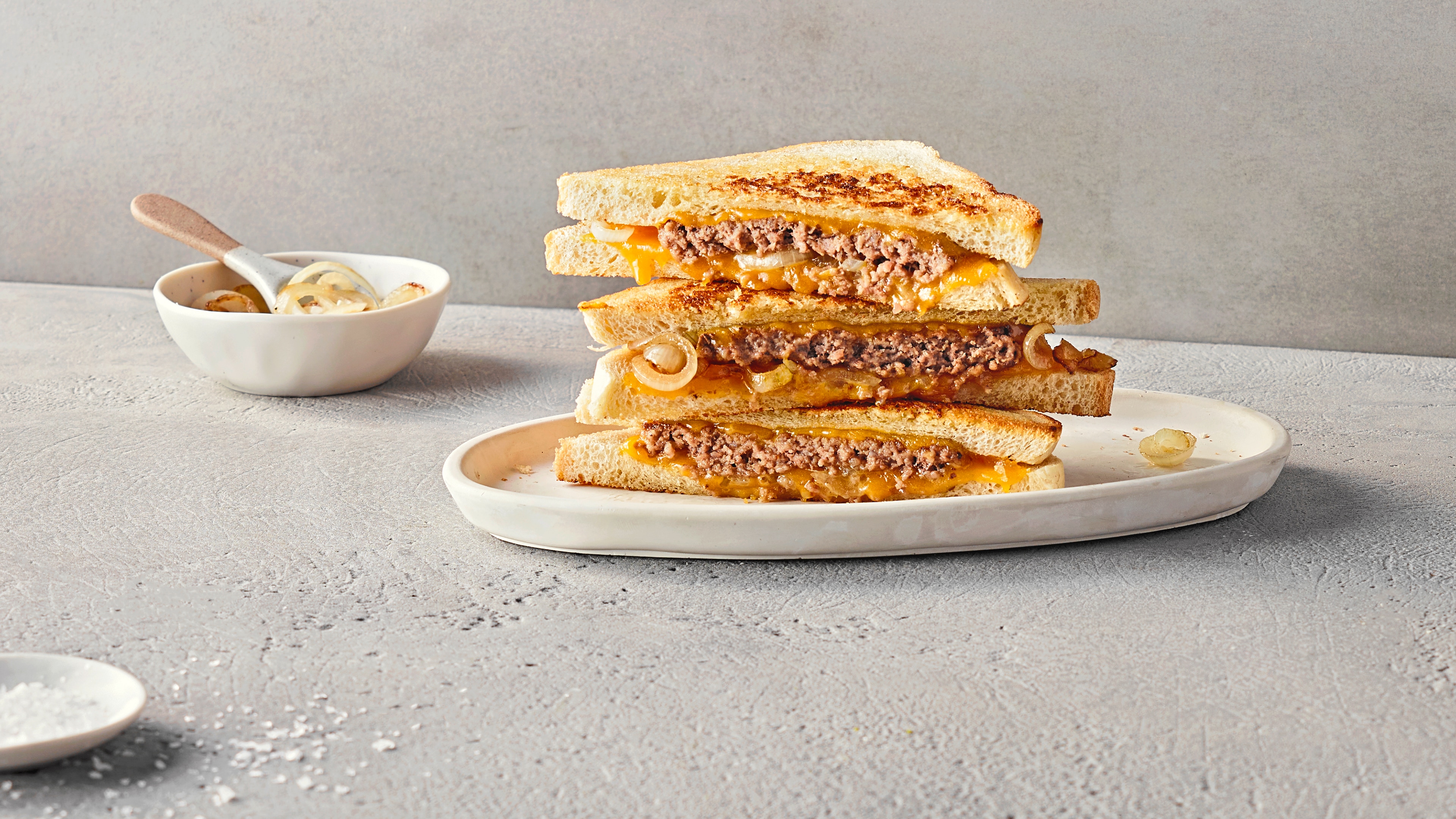 Drei getoastete Sandwichtoast gefüllt mit Hackfleisch, Käse und Zwiebeln auf einem Teller, daneben eine Schale mit Zwiebeln