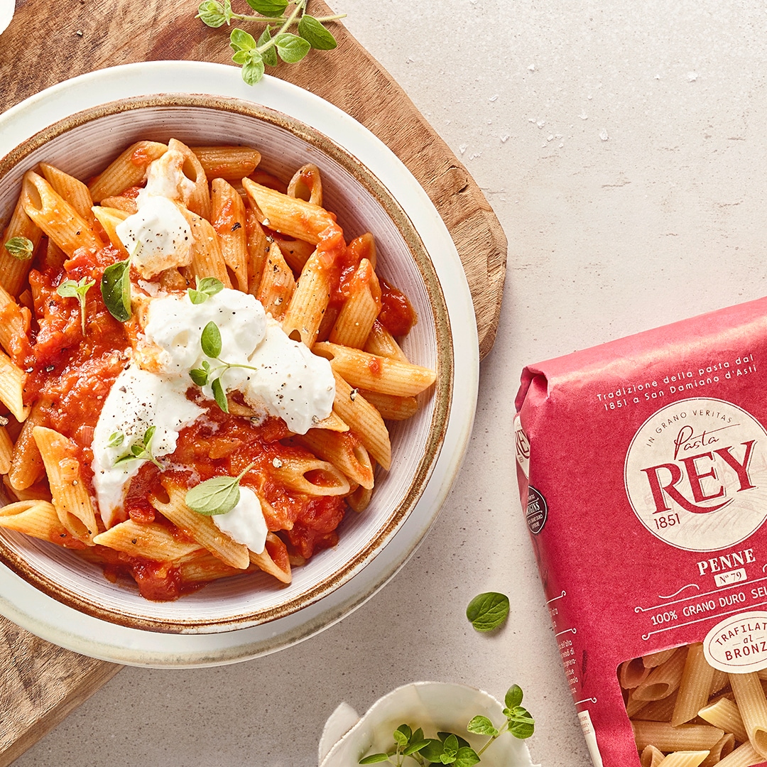 Mit Nudeln, roter Soße und weißem Käse gefüllter Teller auf Holzbrett. Daneben eine Packung Pasta Rey Penne