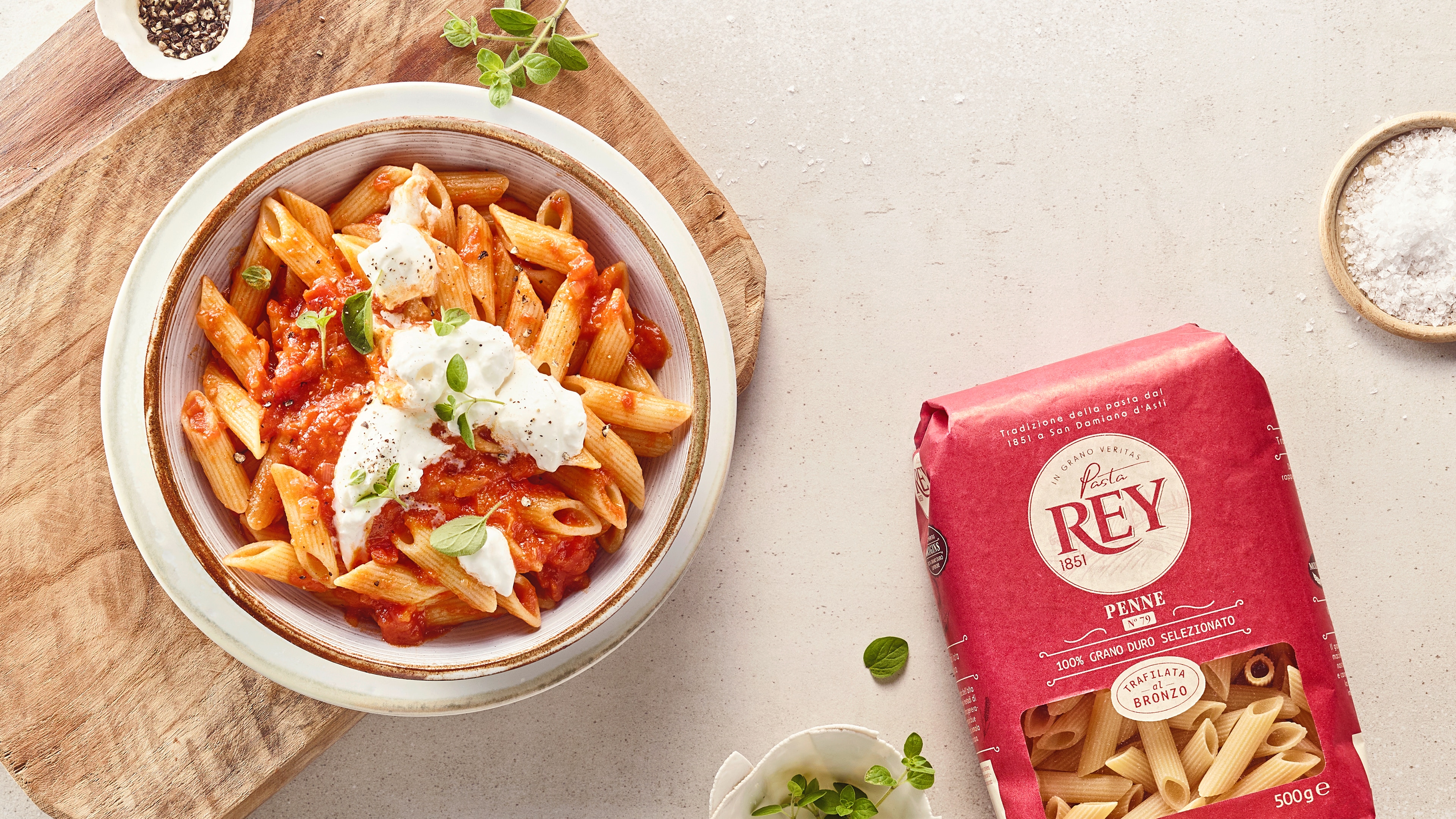 Mit Nudeln, roter Soße und weißem Käse gefüllter Teller auf Holzbrett. Daneben eine Packung Pasta Rey Penne