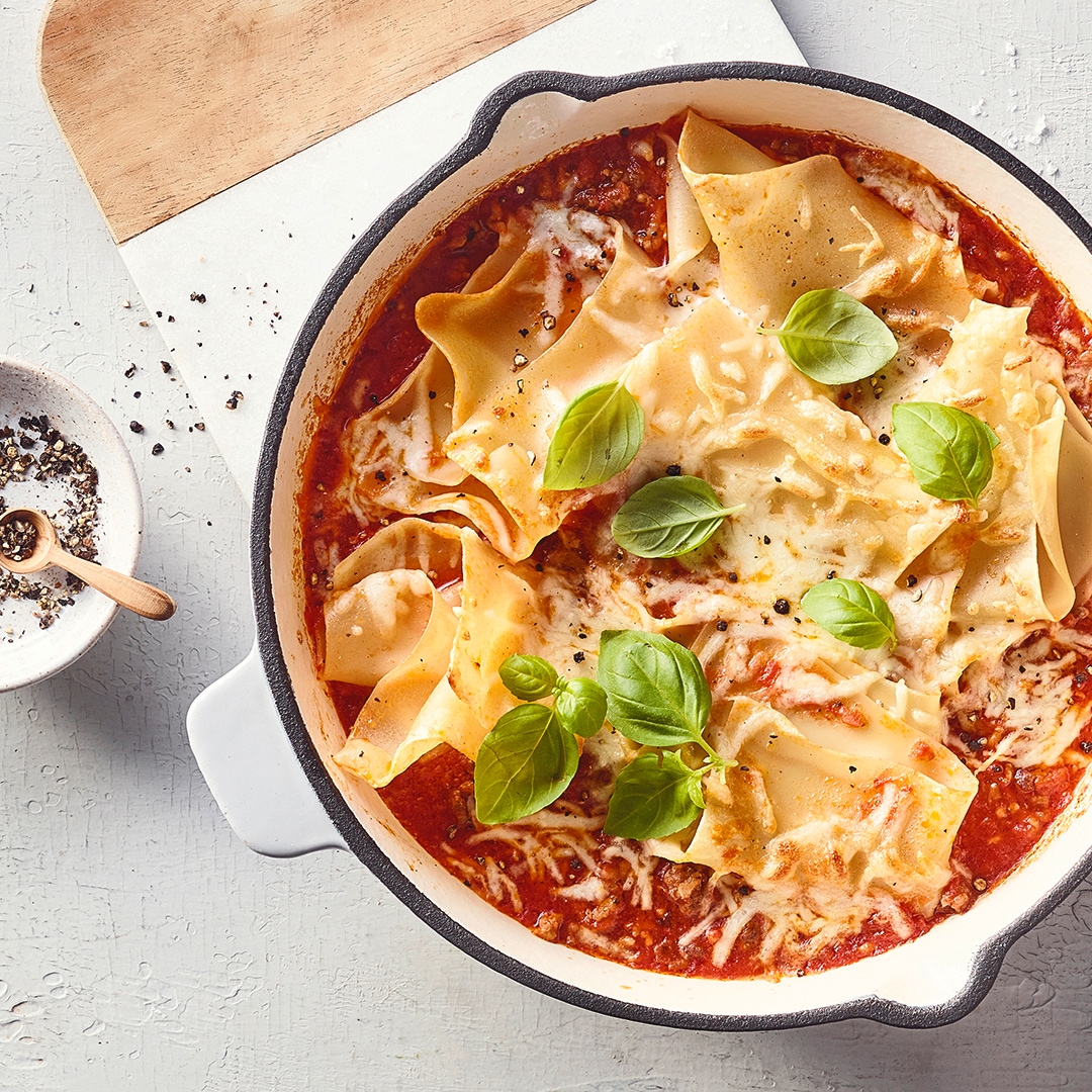 Eine Pfanne mit Lasagneblättern in roter Sauce, bestreut mit geschmolzenem Käse und garniert mit frischen grünen Basilikumblättern