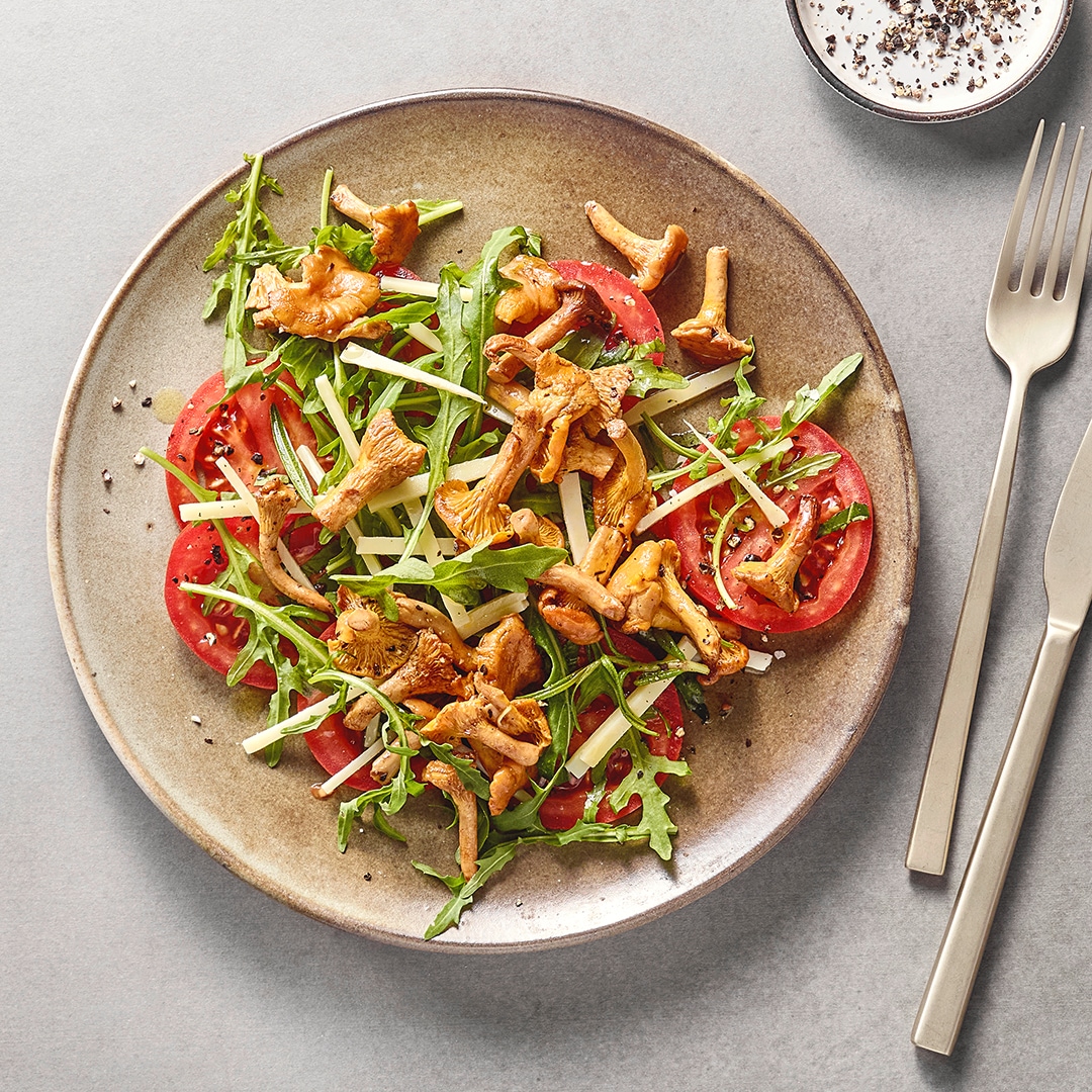 Salat mit Rucola, Tomatenscheiben, Pfifferlingen und gehobeltem Käse auf einem Teller, daneben Besteck.