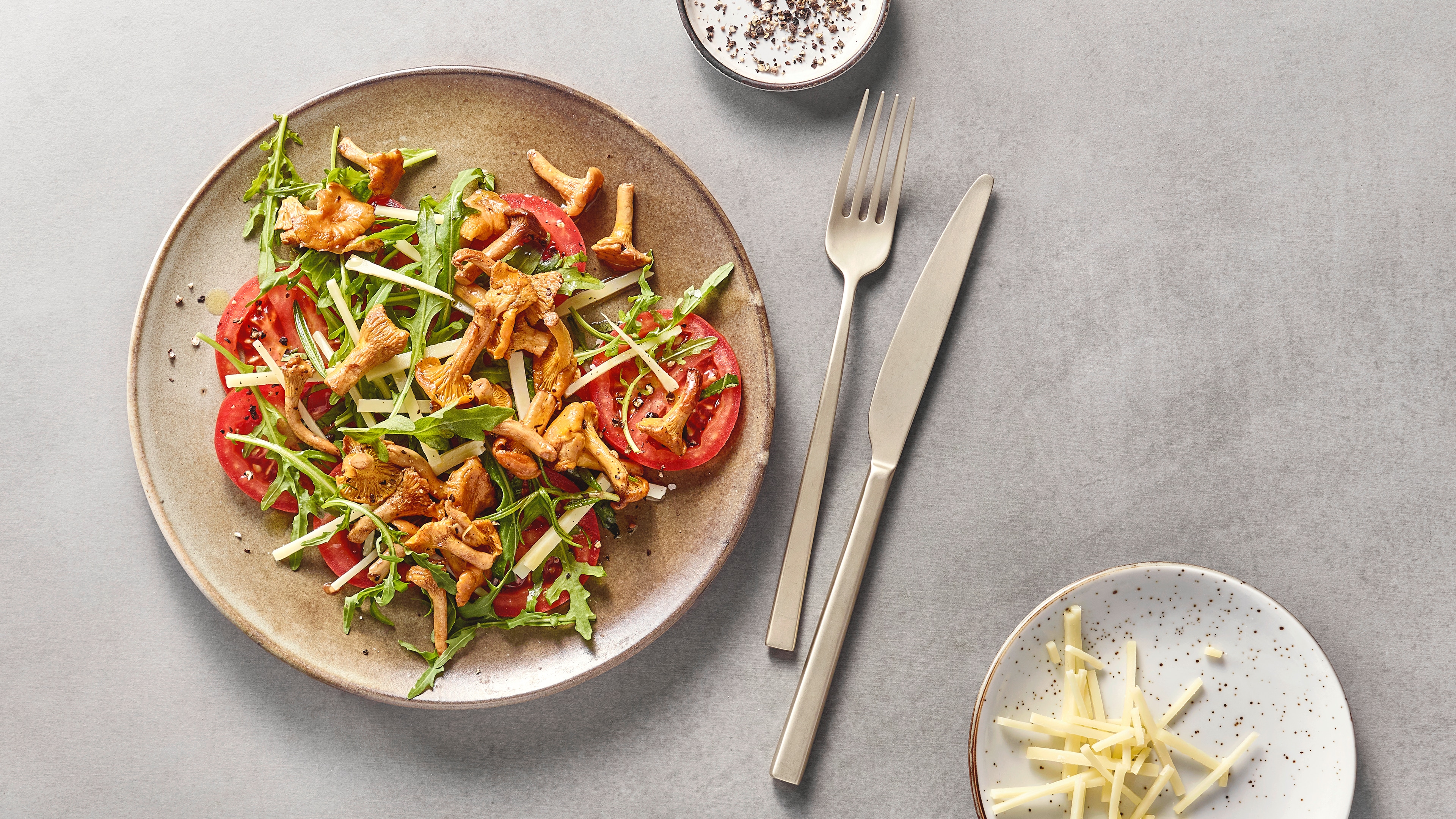 Salat mit Rucola, Tomatenscheiben, Pfifferlingen und gehobeltem Käse auf einem Teller, daneben Besteck und zusätzliche Käsestreifen.