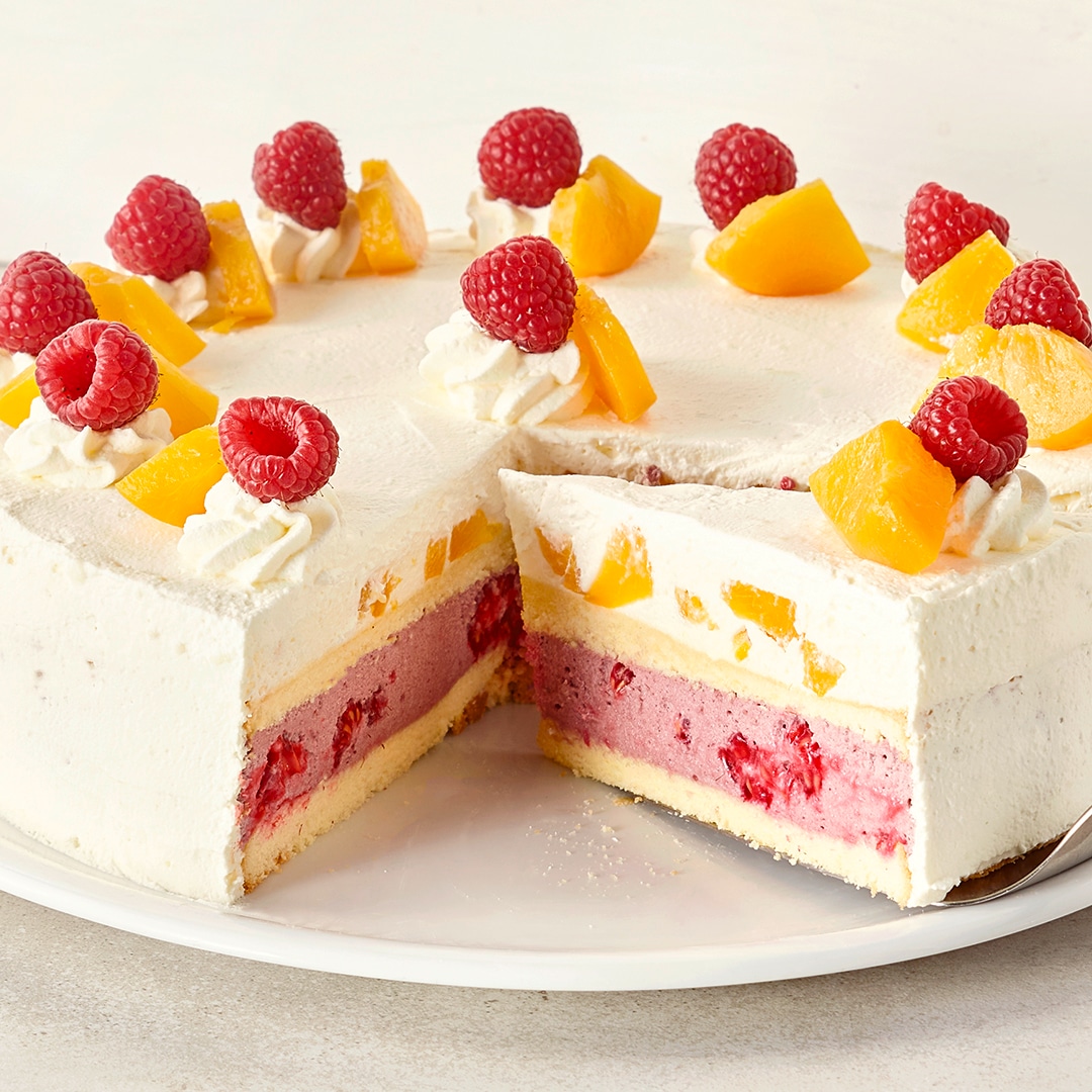 Ein Teller mit einer weißen Torte, gefüllt mit rosa Creme und Früchten, garniert mit Himbeeren und gelben Pfirsichstücken.
