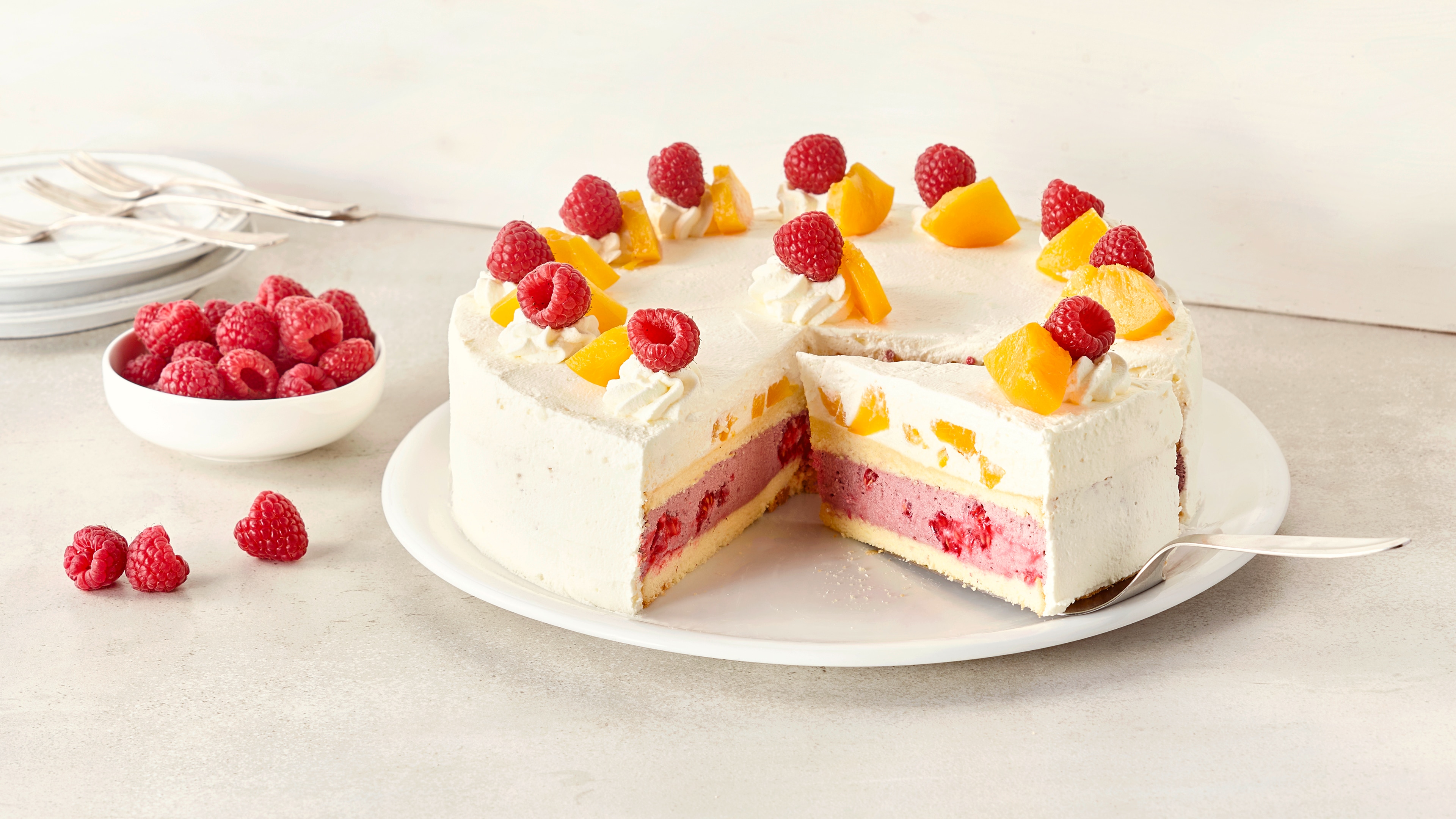 Ein Teller mit einer weißen Torte, gefüllt mit rosa Creme und Früchten, garniert mit Himbeeren und gelben Pfirsichstücken.